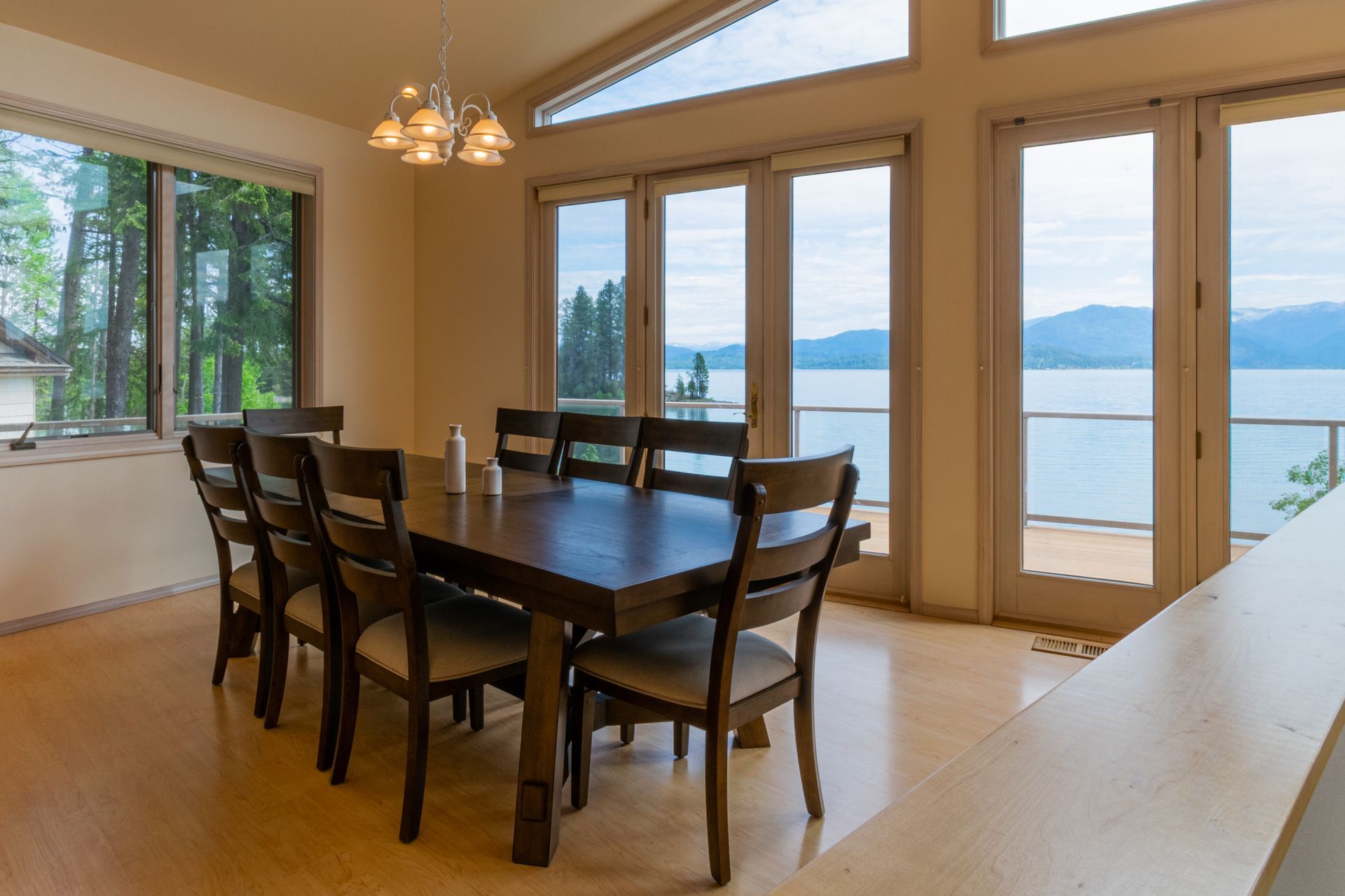 Dining room with large wooden table and chairs, overlooking a lake through expansive windows.