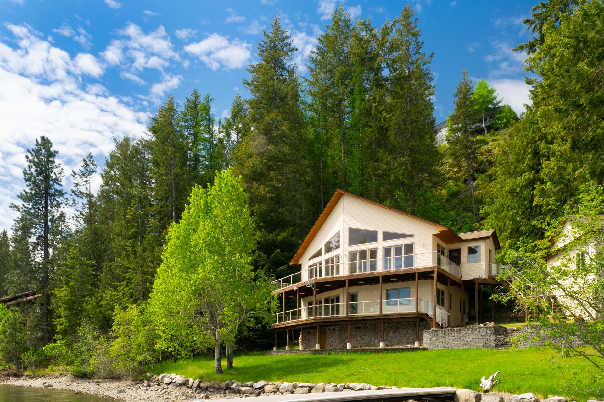 Two-story house with a deck, nestled amongst green trees on a hillside by a lake under a blue sky with clouds.
