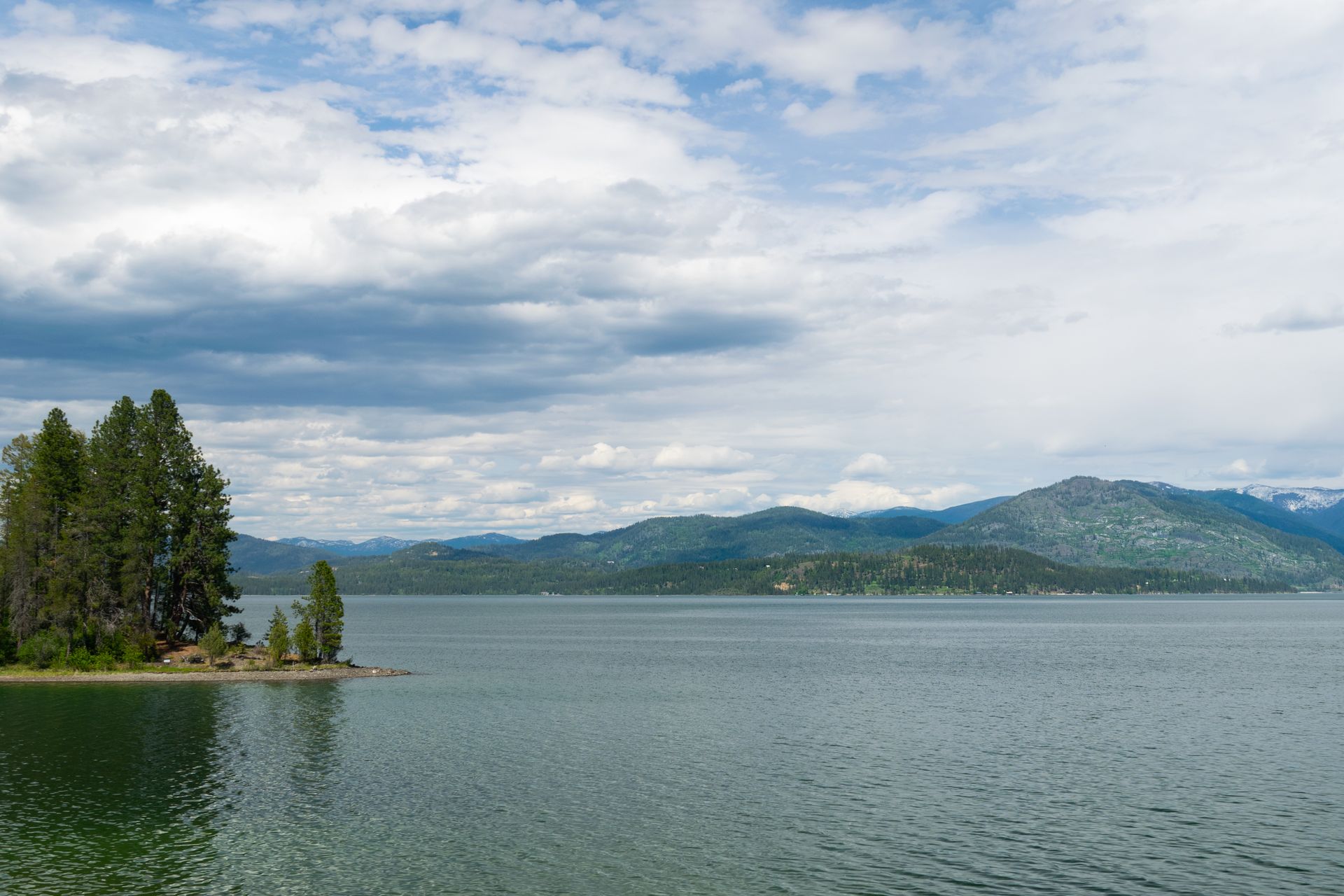 Lake surrounded by mountains under a cloudy sky, with a small island in the foreground featuring tall trees.