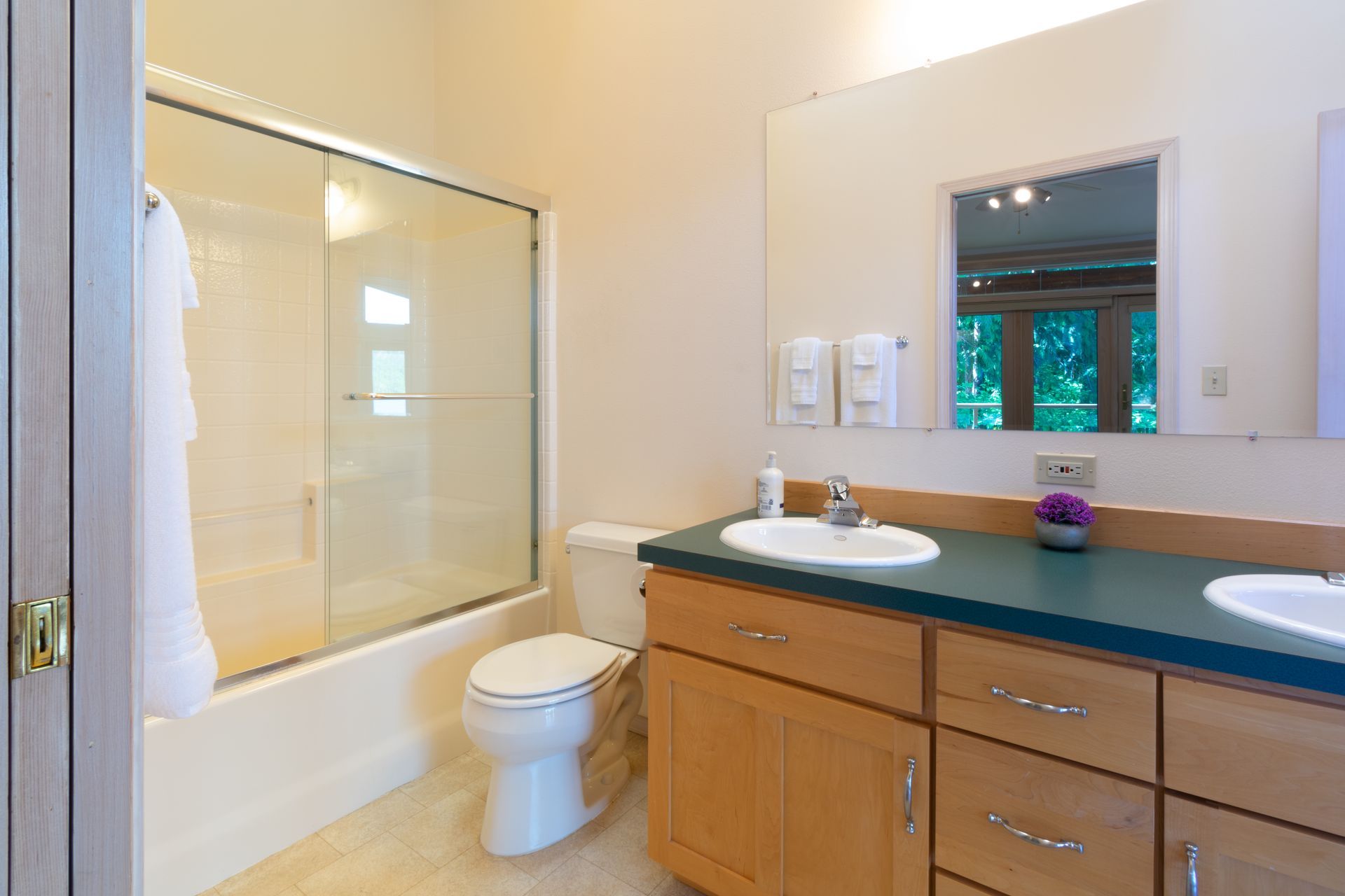 Bathroom with a light beige color scheme, featuring a toilet, double vanity with teal countertop, and a glass shower.