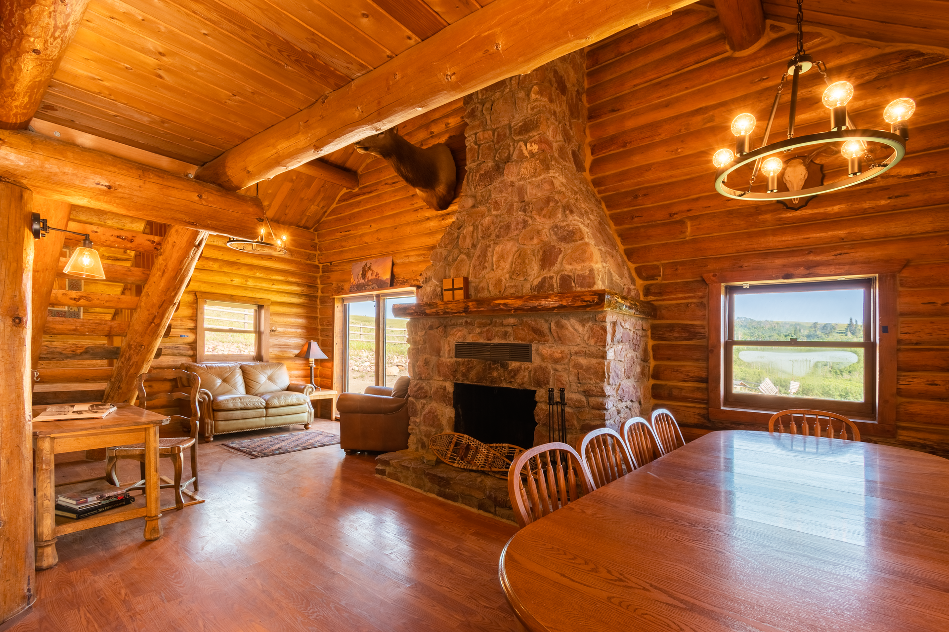 Cozy log cabin interior with stone fireplace, long wooden dining table, and staircase. Sunlight streams in through windows.
