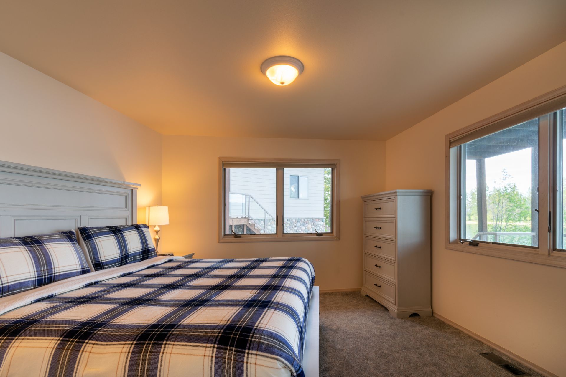 Bedroom with a large bed, a chest of drawers, and windows overlooking a patio. The bed has blue plaid bedding.