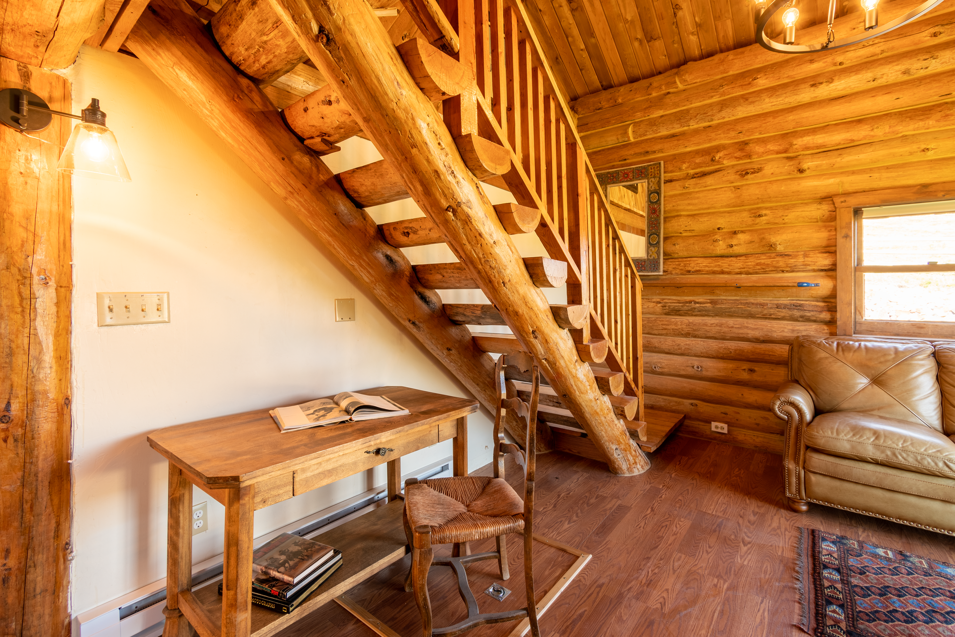 Log cabin interior with wooden staircase, desk, chair, and leather sofa.