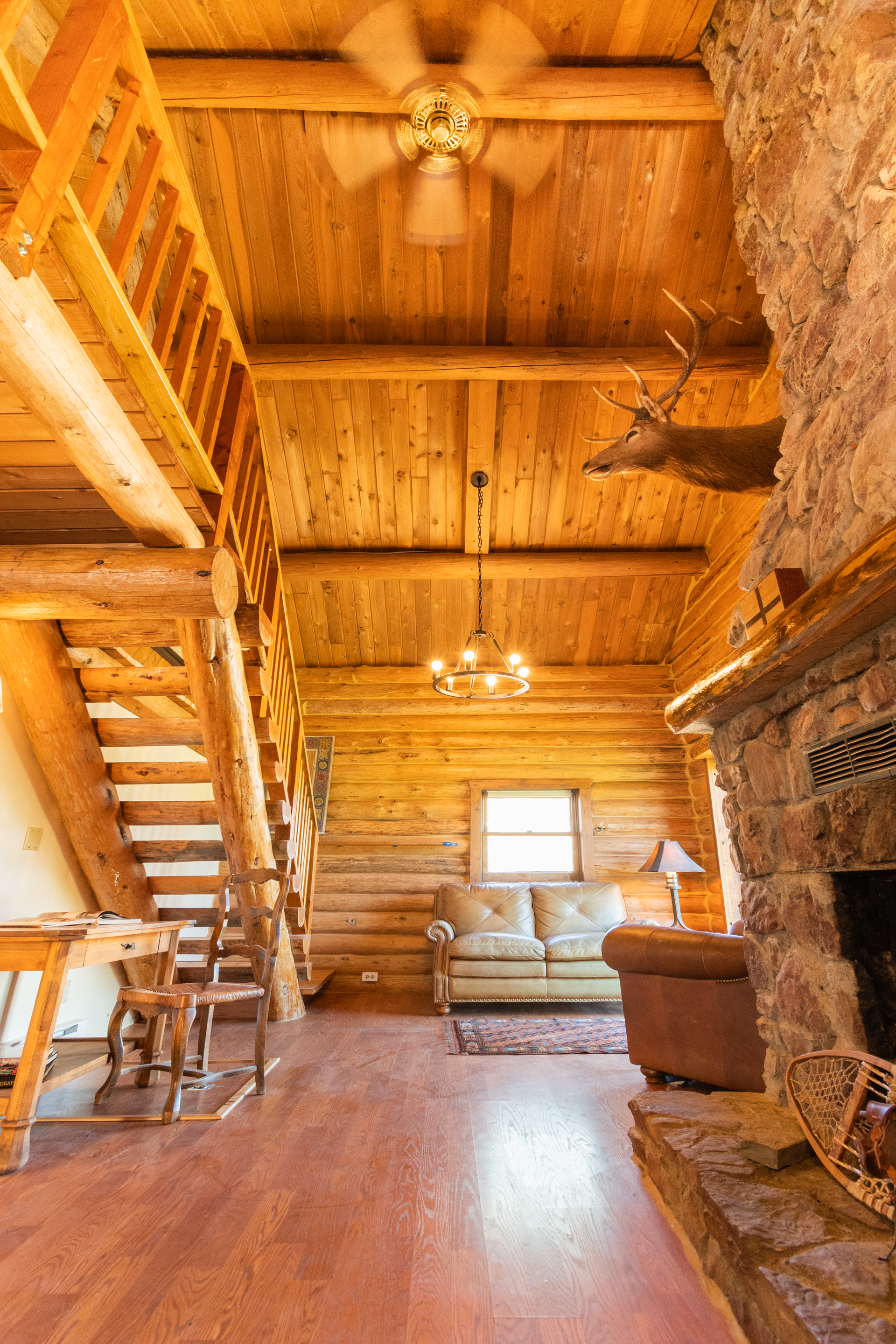 Rustic log cabin interior with wooden floors, stone fireplace, staircase, and a deer head mounted above the fireplace.