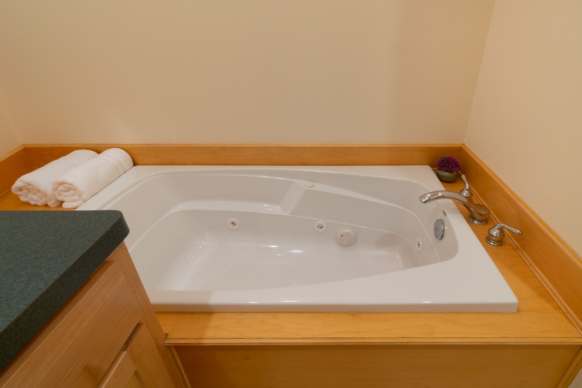 A built-in white jacuzzi tub surrounded by wooden trim, with rolled towels on the counter and beige walls.
