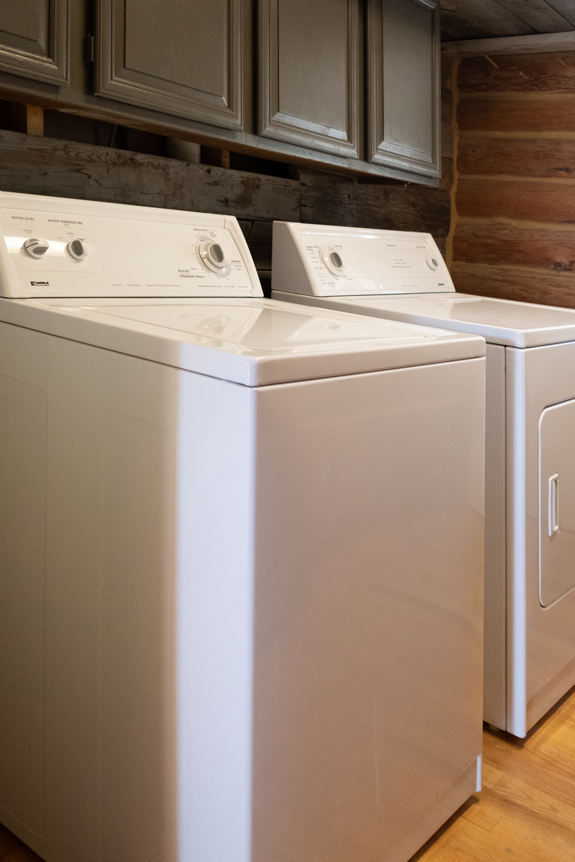 A washing machine and dryer set in a laundry room, with upper cabinets and wood paneling. Both appliances are white.