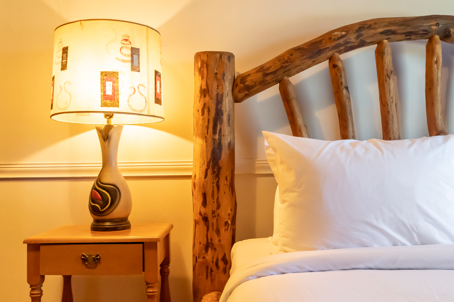 Bedroom with a rustic wood headboard, nightstand, lamp, and a white pillow on the bed.