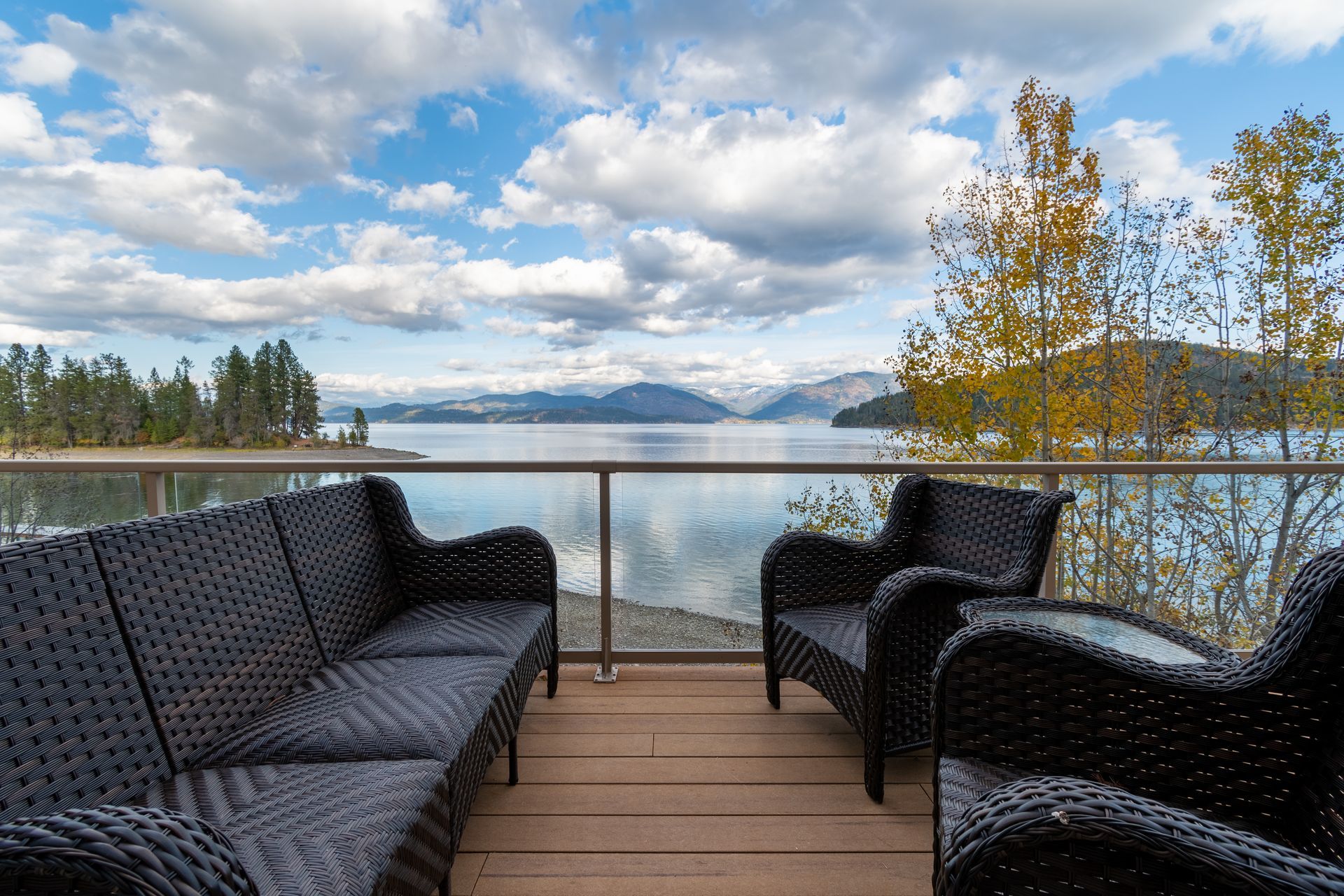 A waterfront patio with black wicker furniture overlooks a lake and distant mountains under a cloudy sky.