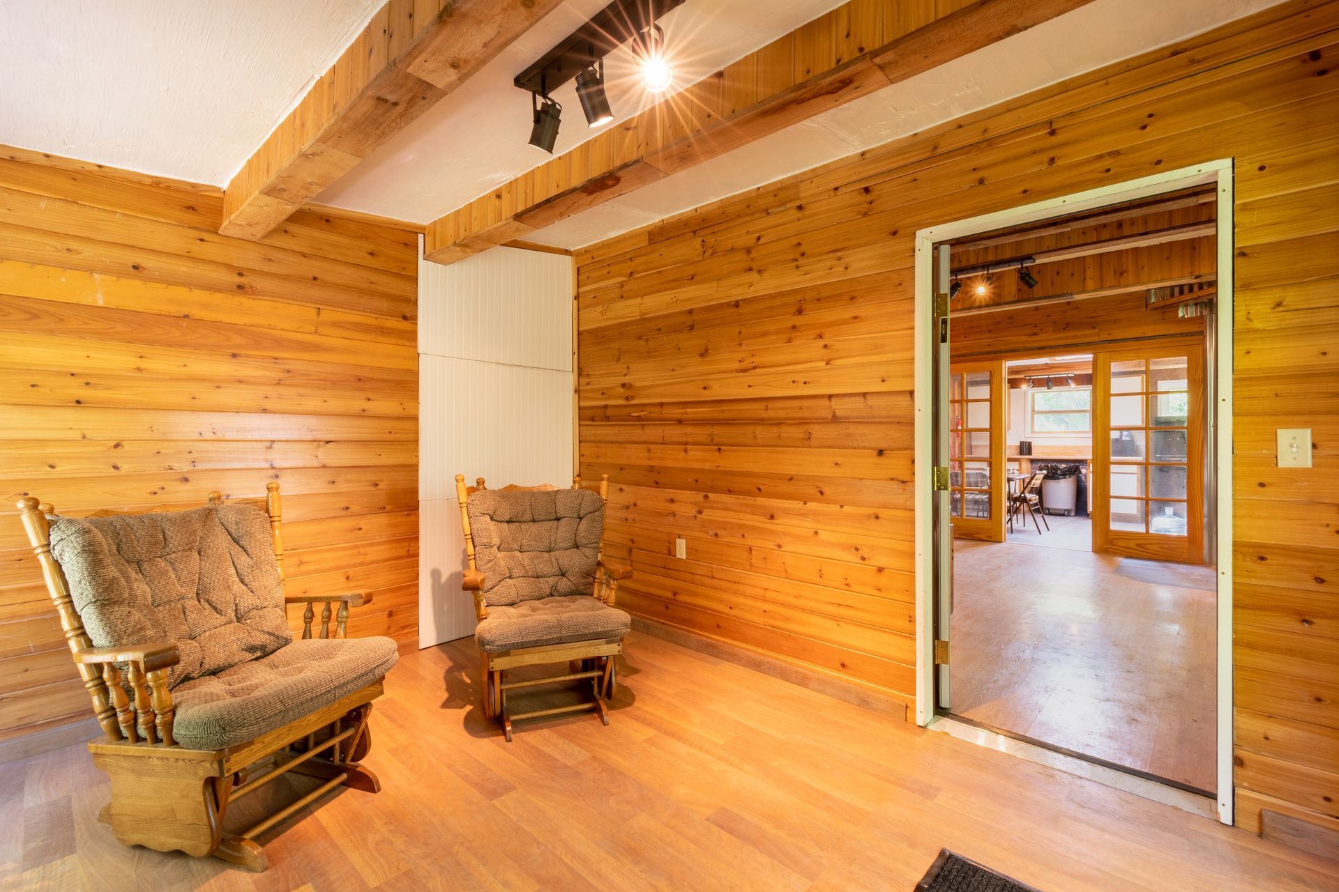 Cozy room with wood-paneled walls and ceiling, two rocking chairs, and a doorway to another room.
