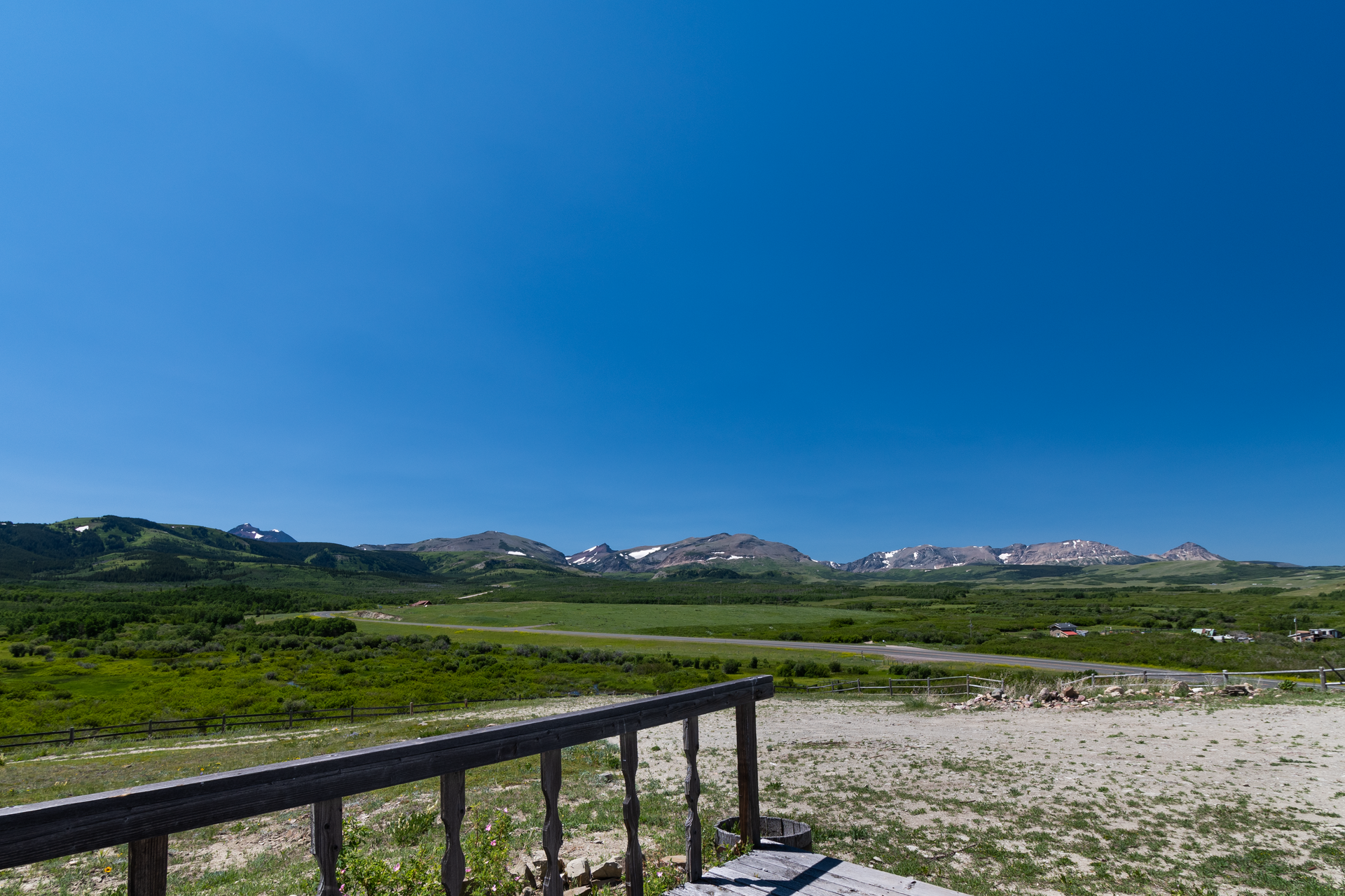 View from a wooden railing across a grassy valley towards a range of mountains under a bright blue sky.