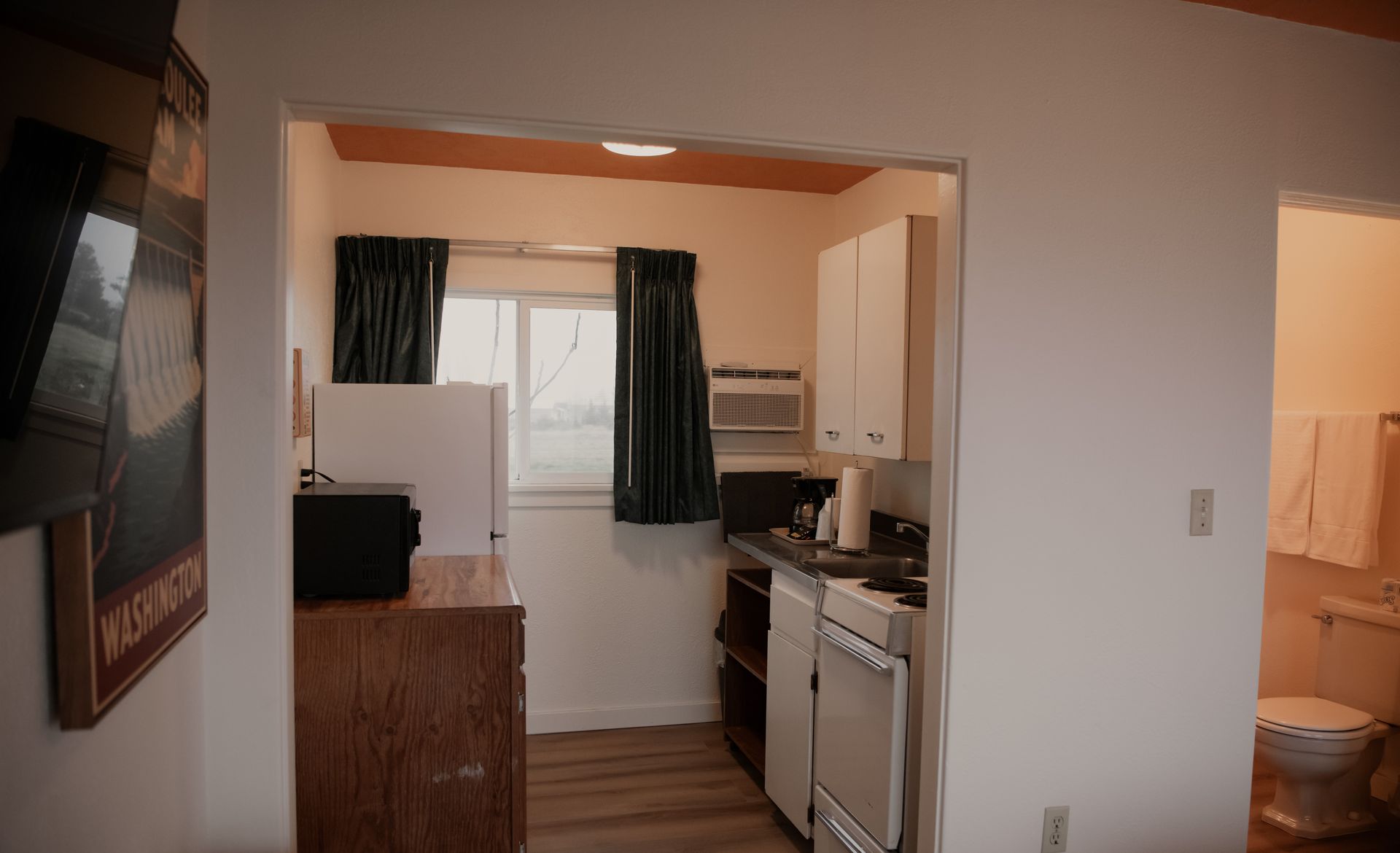 Small kitchen with white cabinets, appliances, and a window. An adjacent room includes a bathroom.