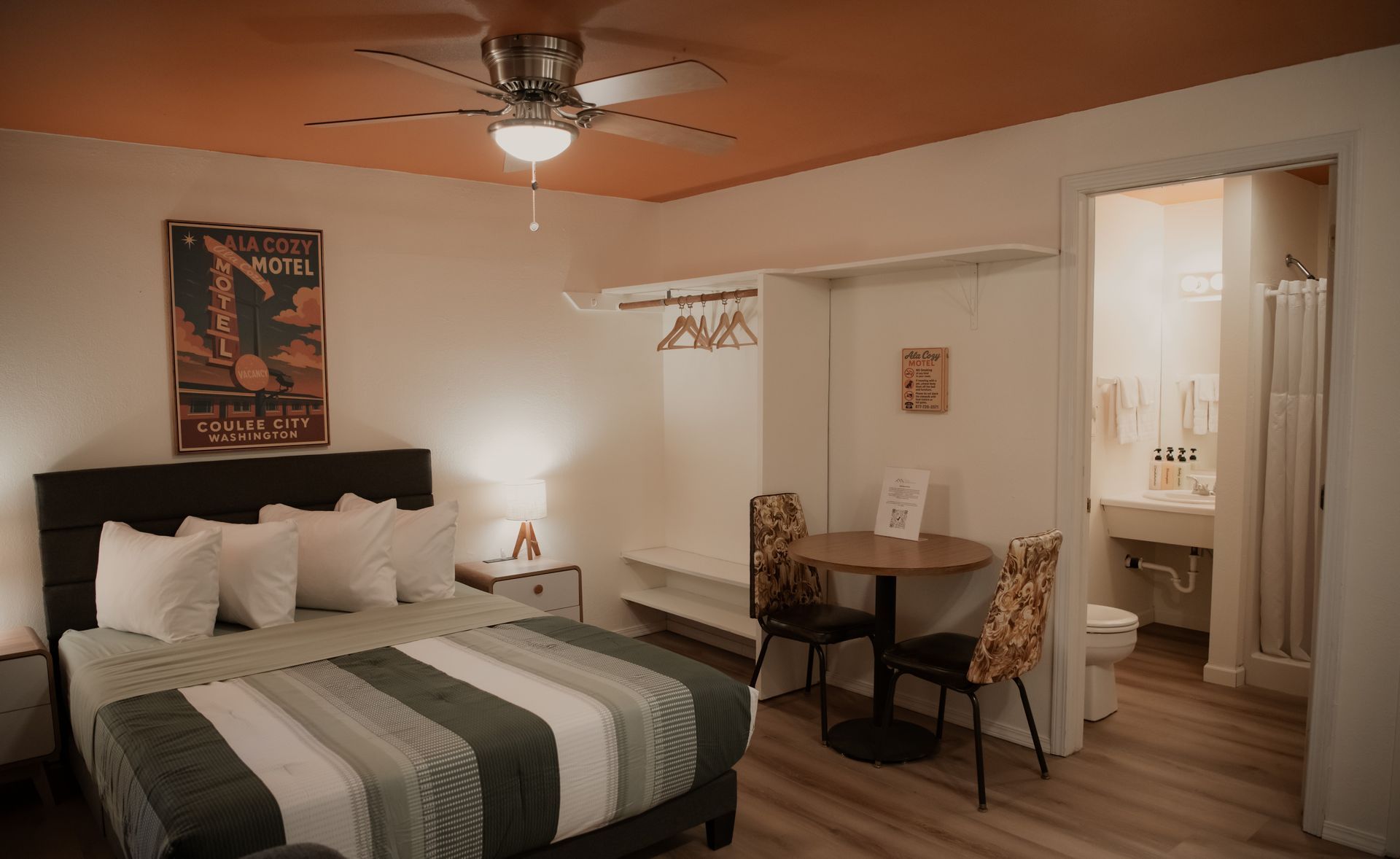 Bedroom interior with bed, table, and bathroom. Orange ceiling, neutral walls, and light wooden floor.