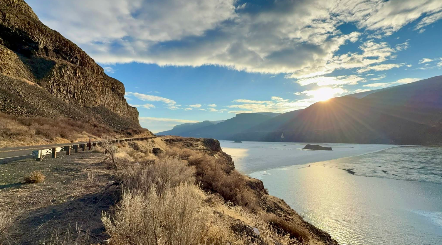 A scenic view of a road and a river bordered by cliffs, with the sun shining through clouds.