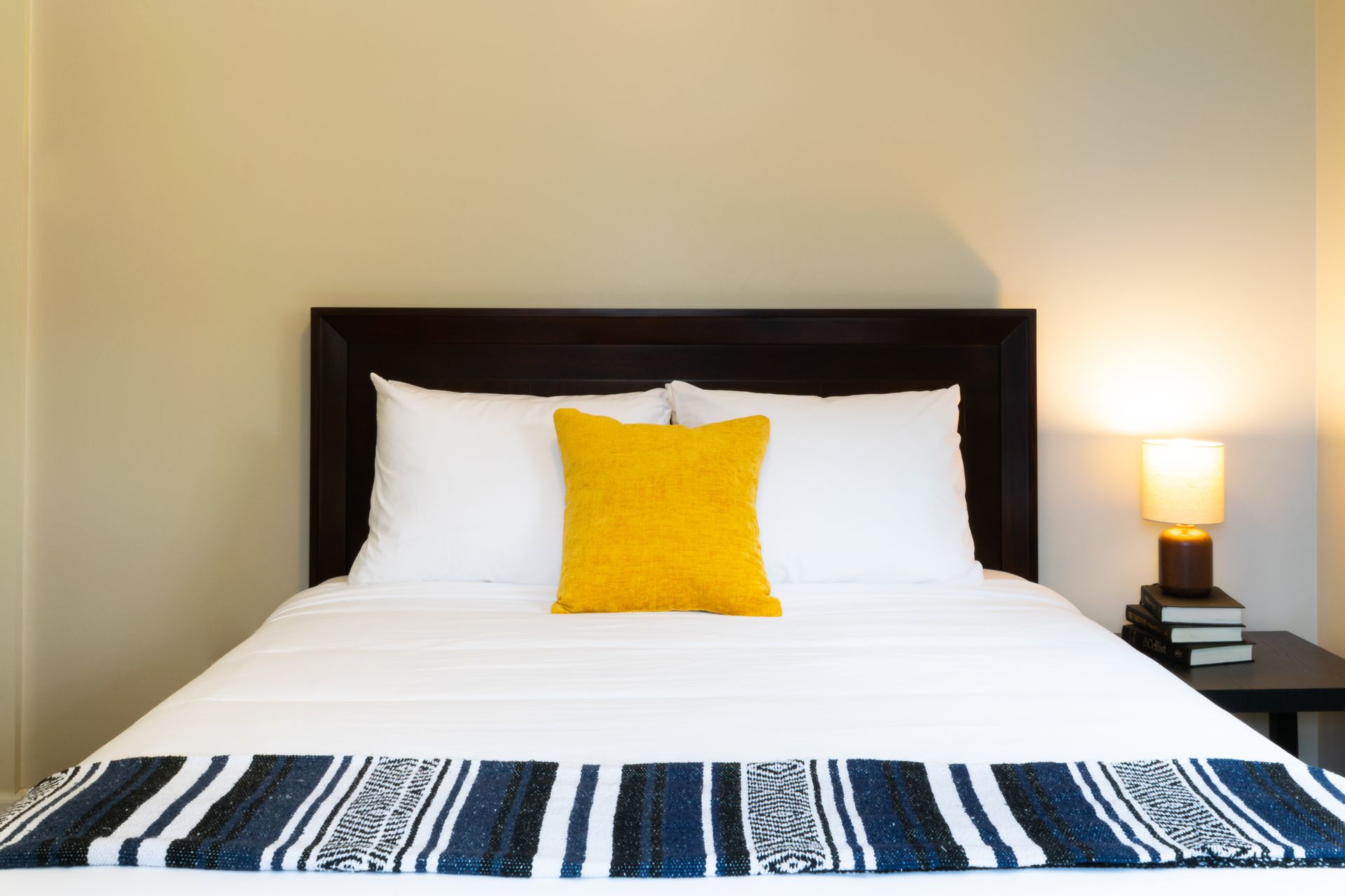 A bed with white sheets, a yellow pillow, and a blue and white striped throw. A lamp sits on a nightstand.