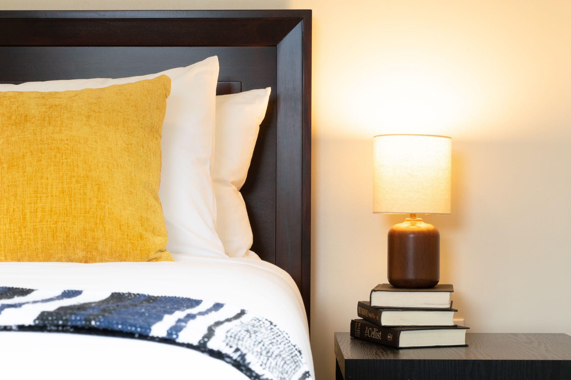 A bed with a dark headboard, a yellow pillow, and a bedside table with a lamp and stacked books.