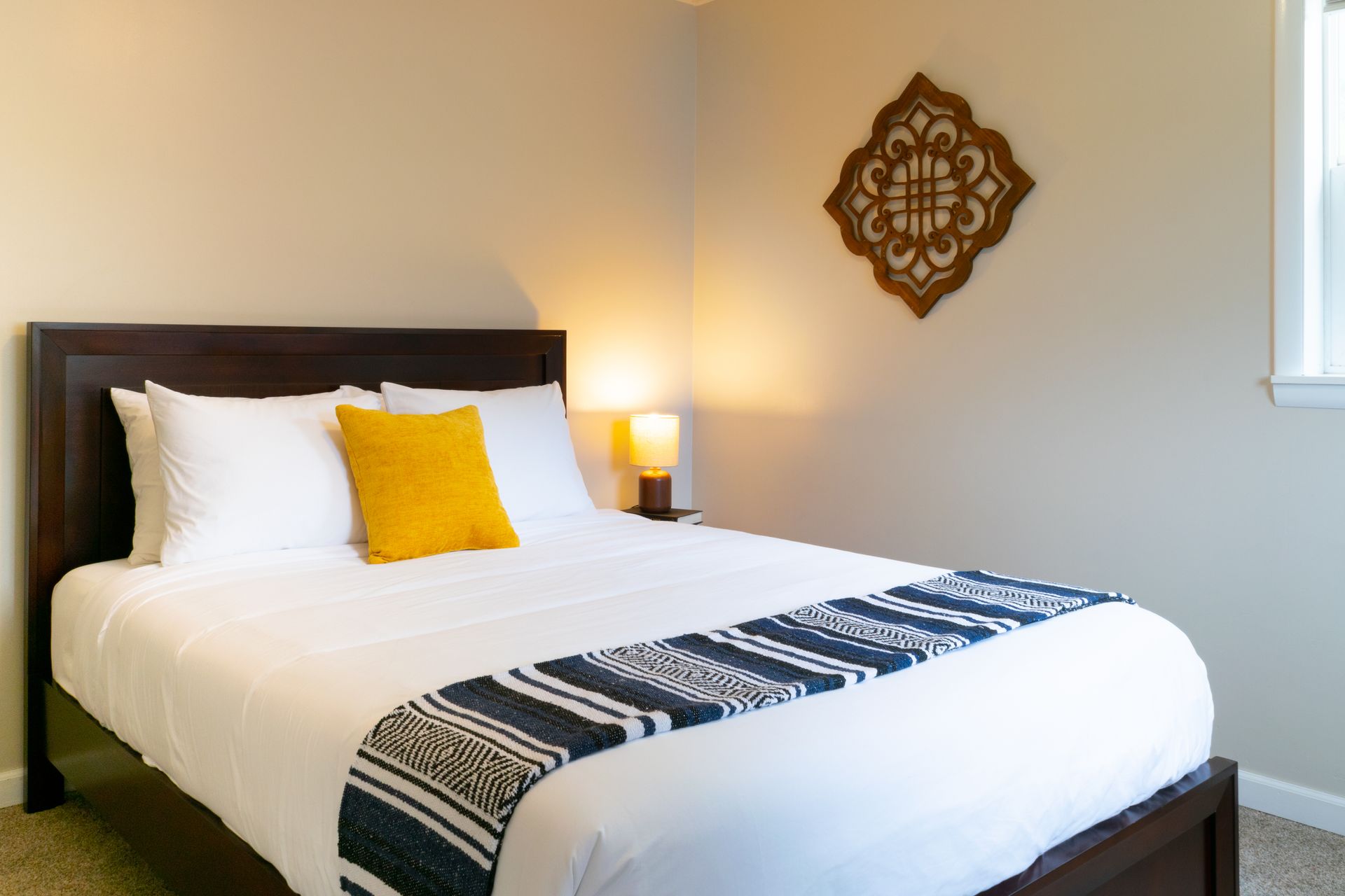 A well-made bed with white linens, a yellow pillow, and a blue and white runner. A wooden decorative piece hangs above.