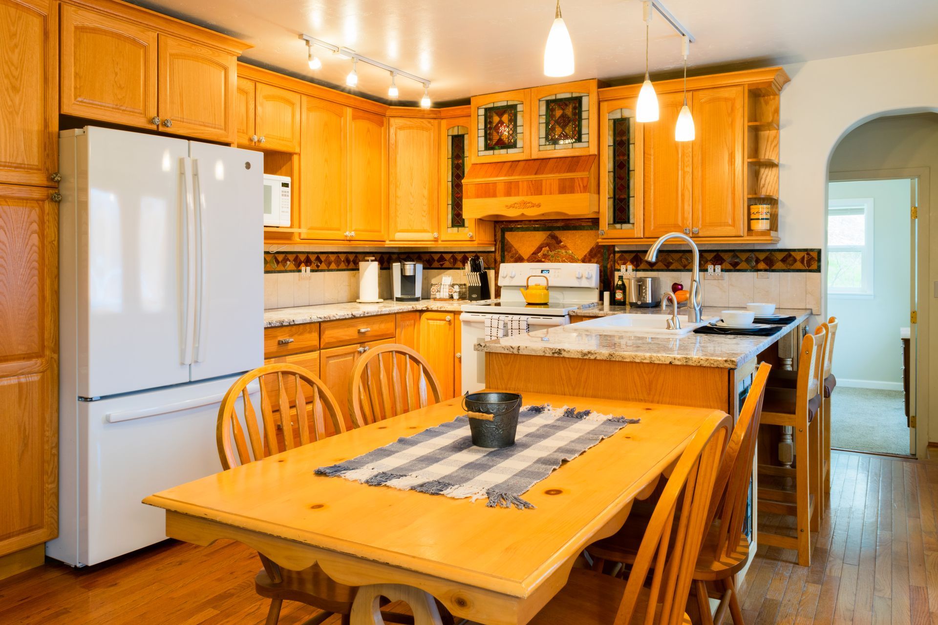 A well-lit kitchen with light wood cabinets, an island, and a dining table with chairs. A white refrigerator is on the left.