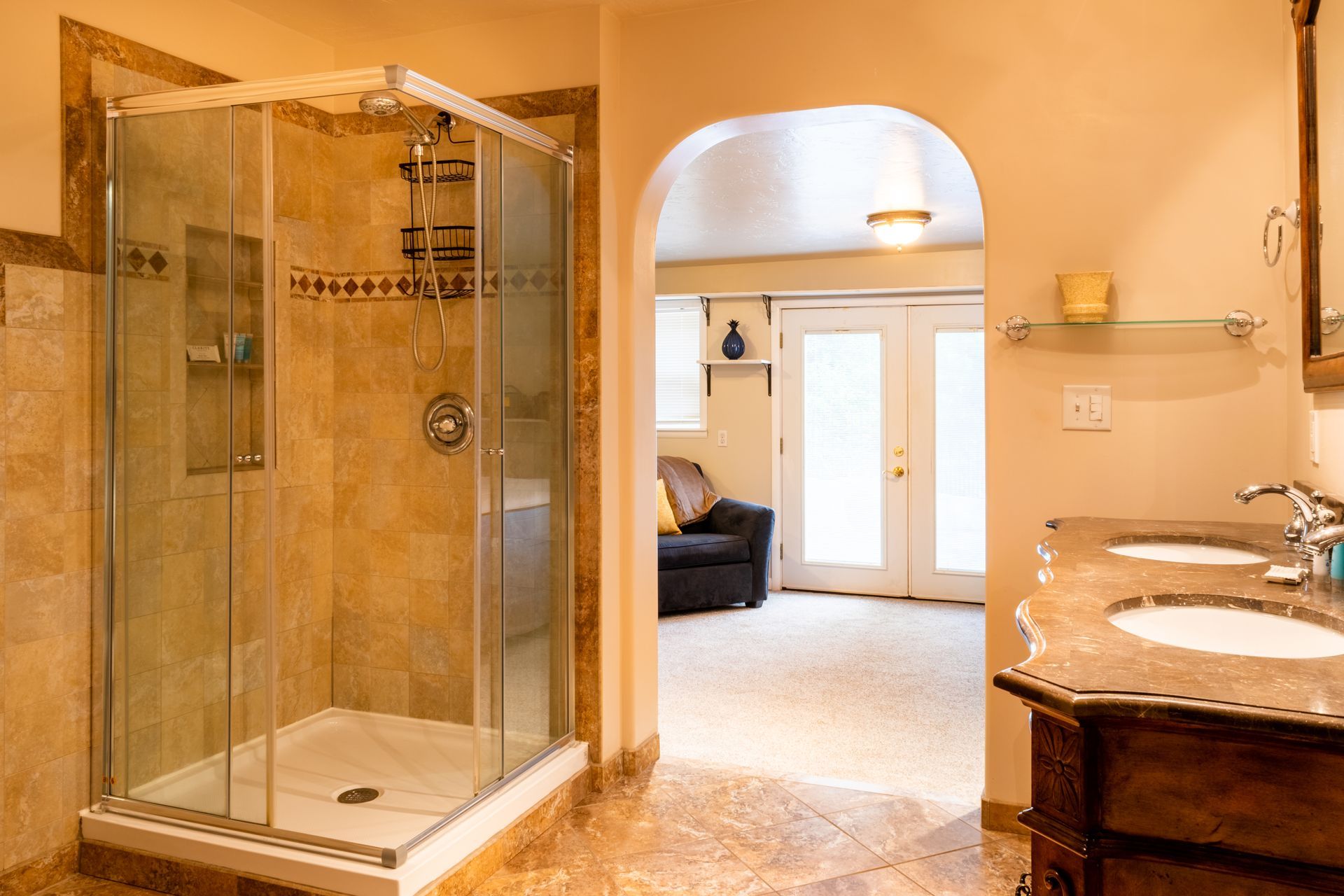 Bathroom with a tiled shower, vanity with two sinks, and an archway leading to a seating area and doors.