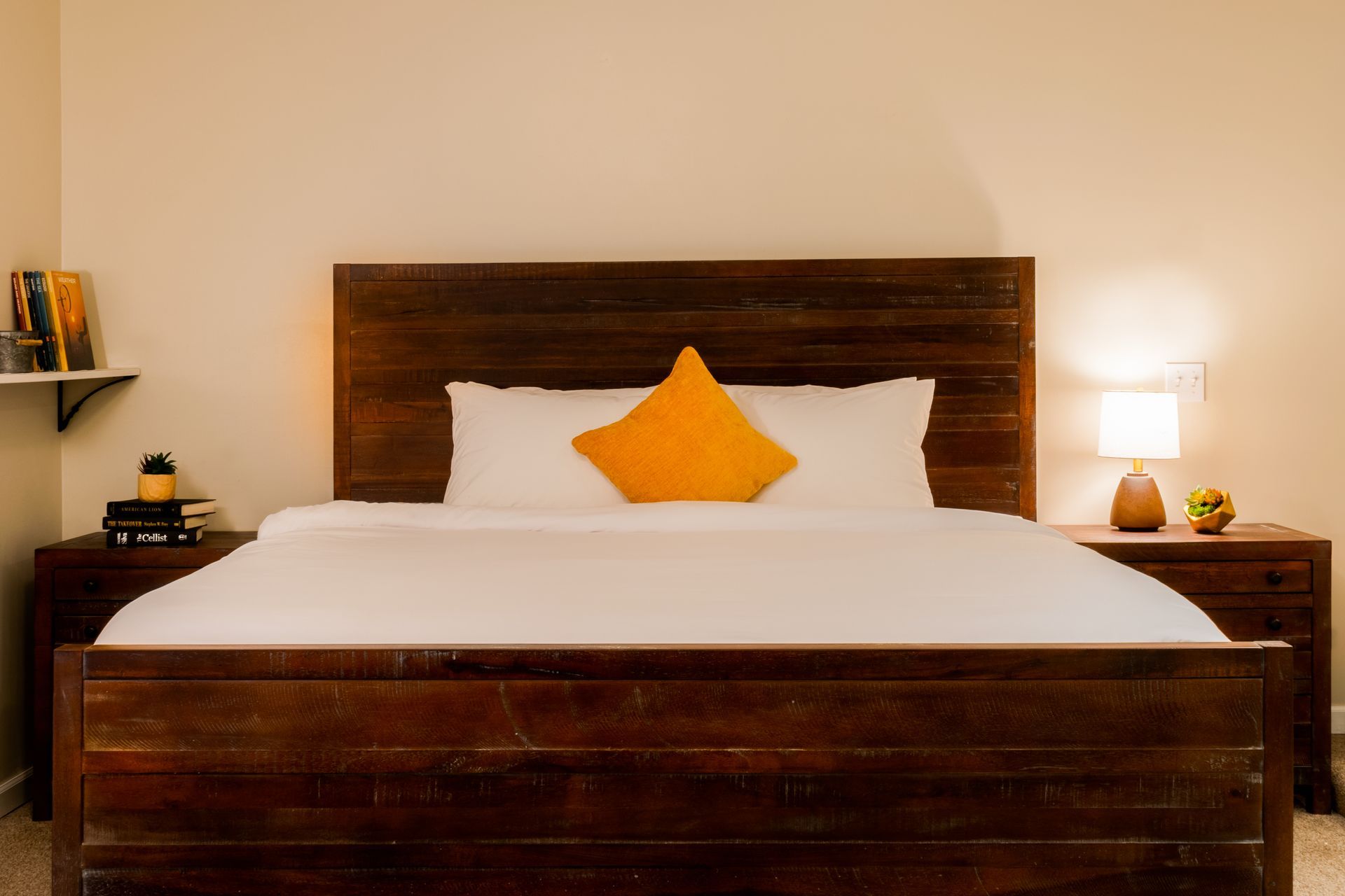 A cozy bedroom with a large wooden bed, white linens, and a yellow pillow. Two nightstands flank the bed, each with a lamp.