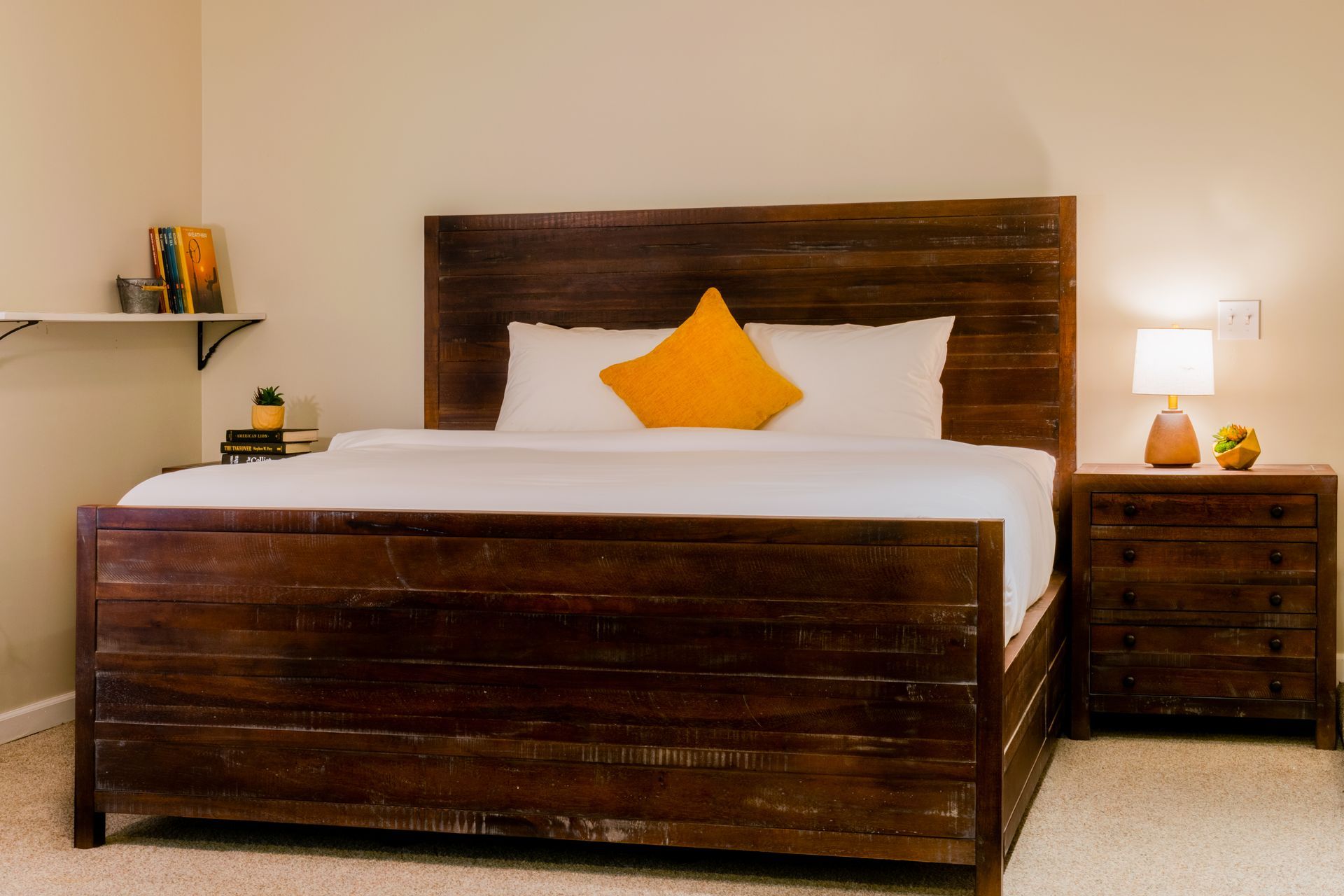 A bedroom with a wooden bed, nightstand, and a shelf. An orange pillow sits on the bed.