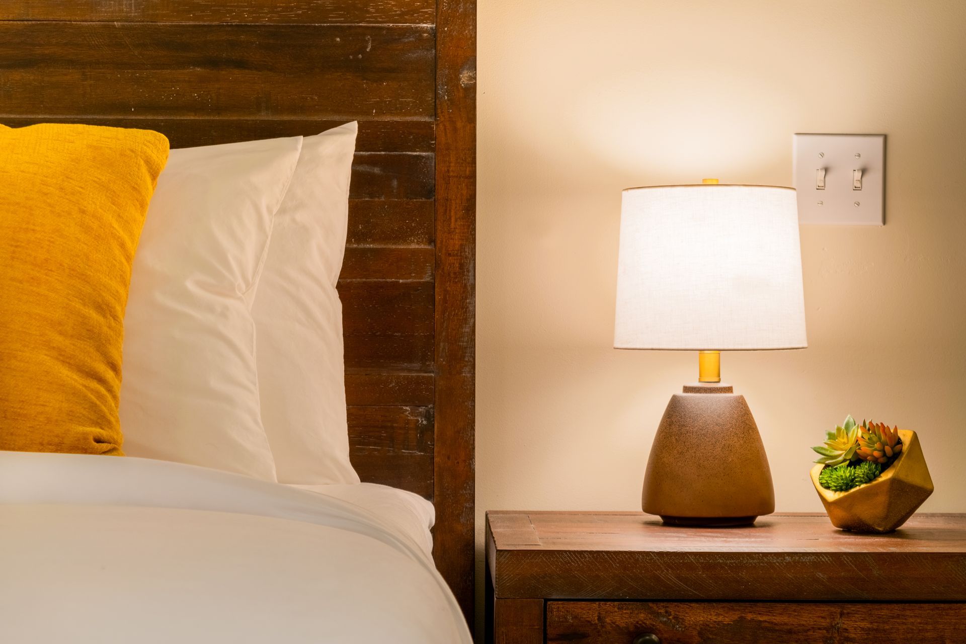 Bedside table with lamp, pillows, and small plant. A warm, inviting bedroom setting with wood tones and yellow accents.