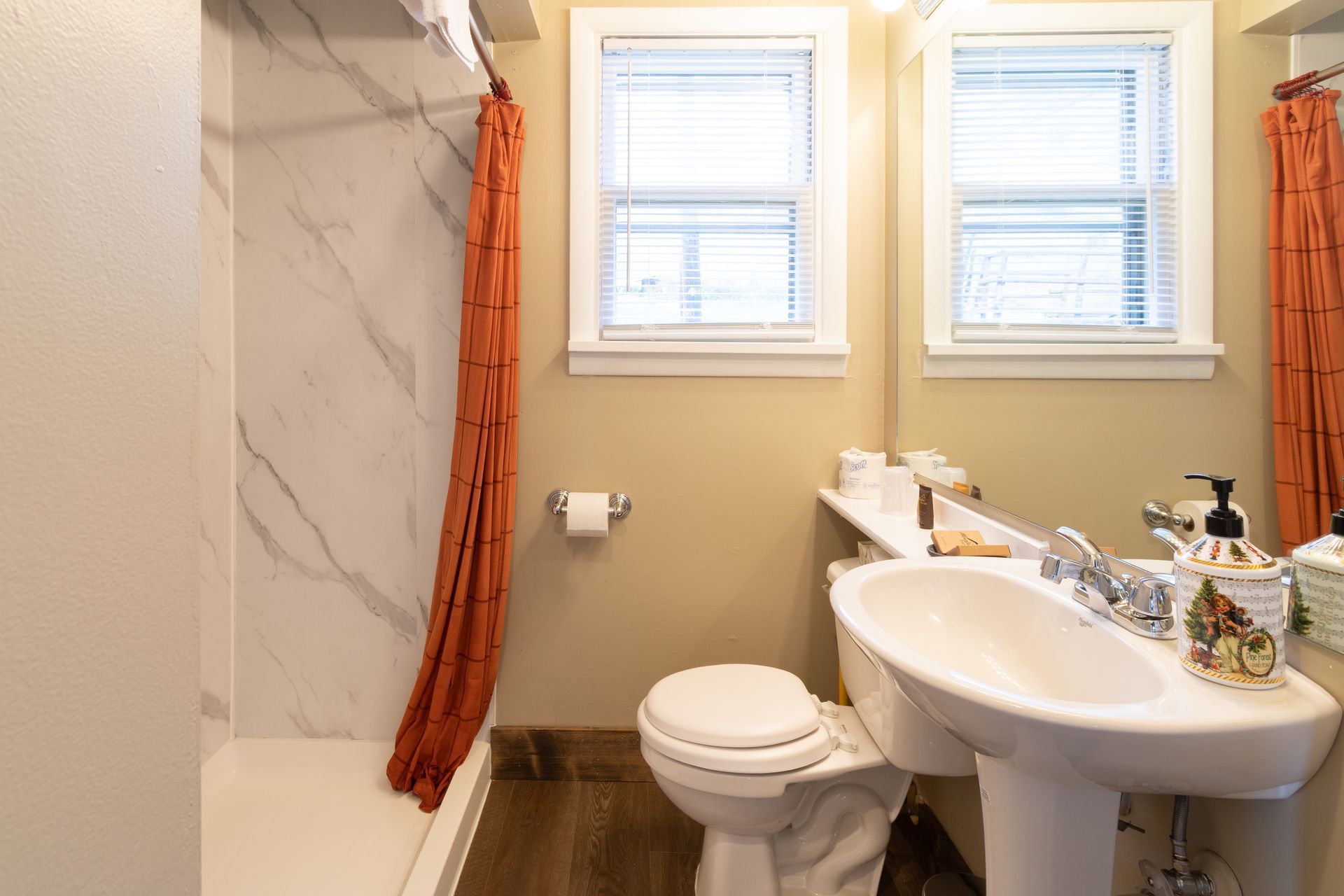 A bathroom featuring a white pedestal sink, toilet, and shower with an orange curtain. Two windows provide natural light.