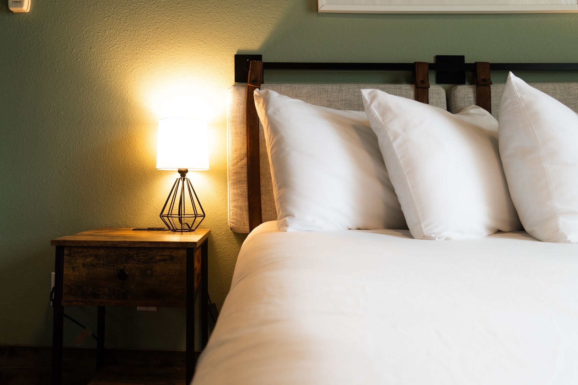 Cozy bedroom scene with a lit bedside lamp on a nightstand next to a bed with white linens and pillows.