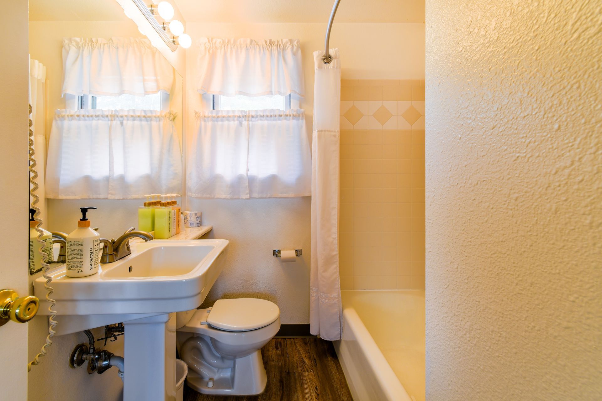 A small bathroom with a pedestal sink, toilet, and bathtub. Soft white curtains frame the windows. Light, textured walls.
