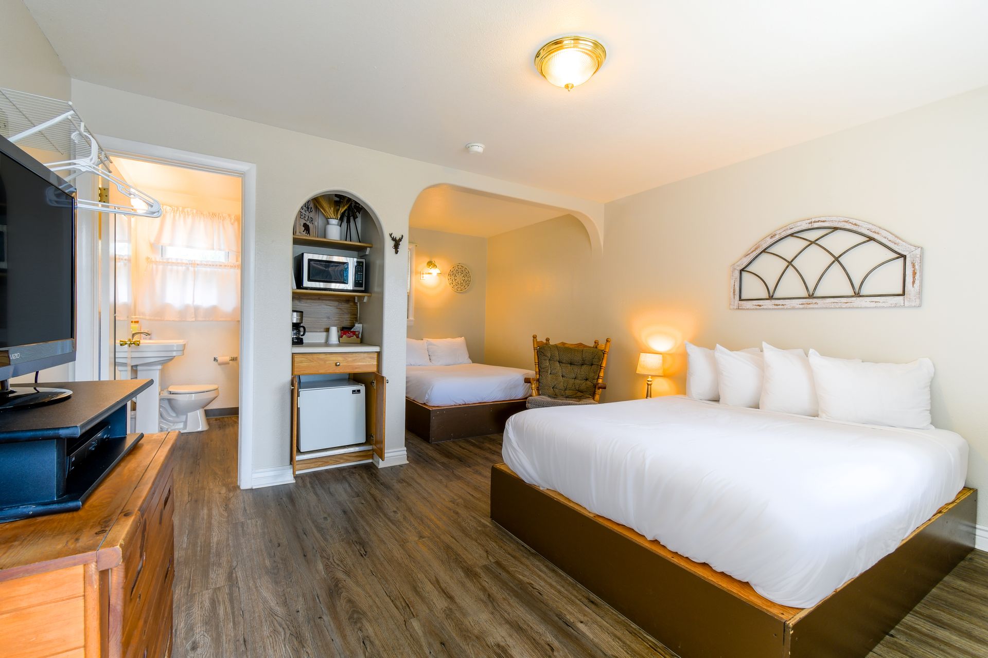 A hotel room with a king-size bed, a smaller bed, and a kitchenette, with an open doorway to a bathroom.