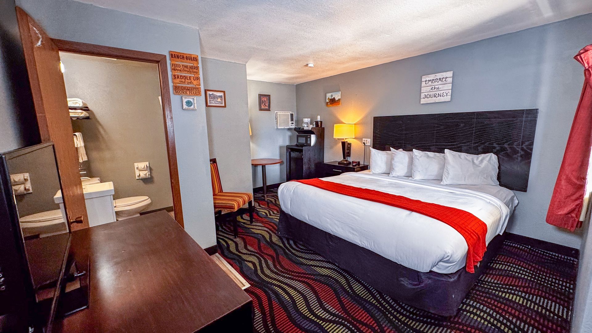 A motel room with a bed, dresser, and a small table. The room has a neutral color palette with green accents.