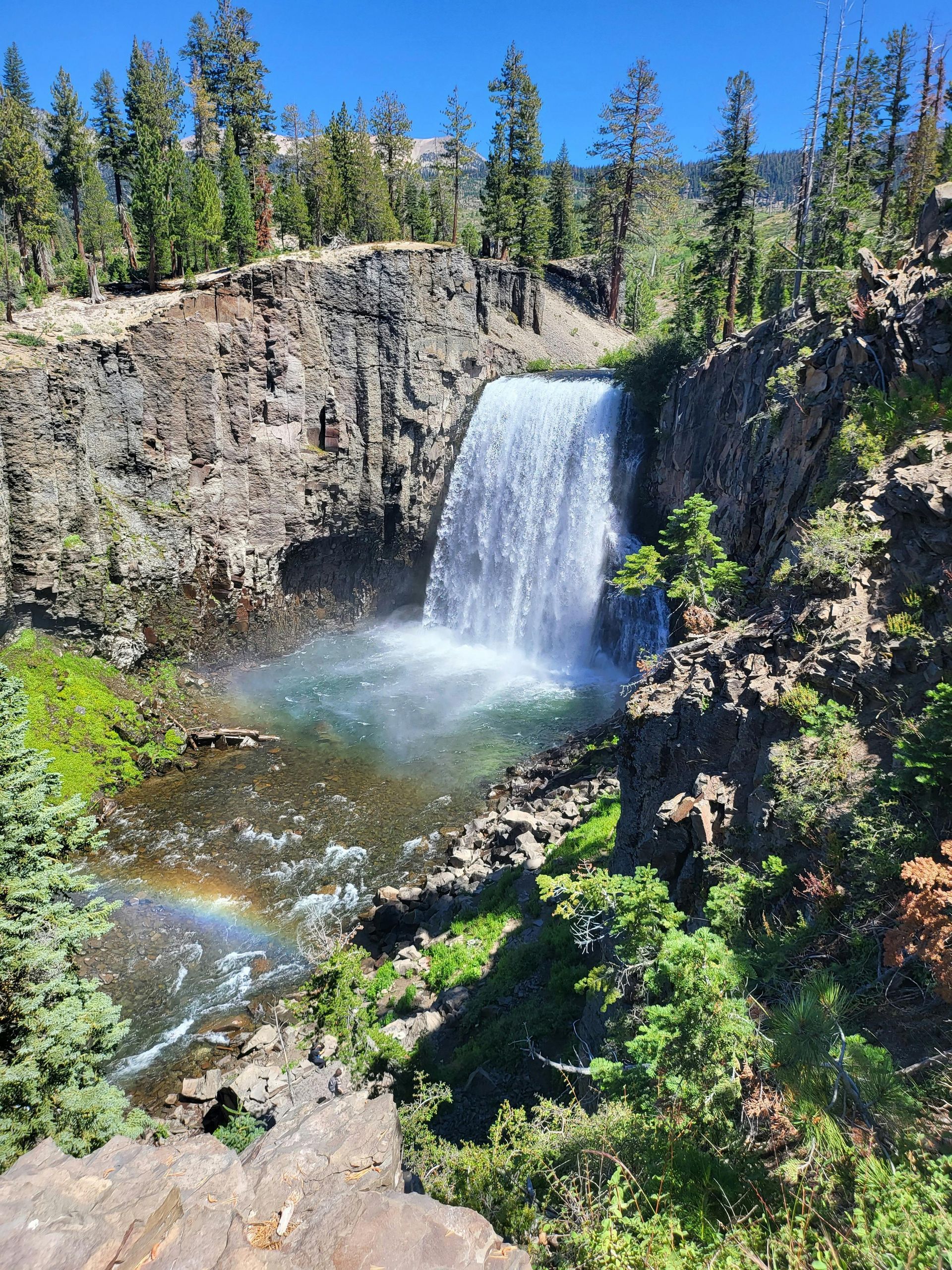 Waterfall cascading into a pool, creating a rainbow. Rocky cliffs surround it, with green trees under a blue sky.