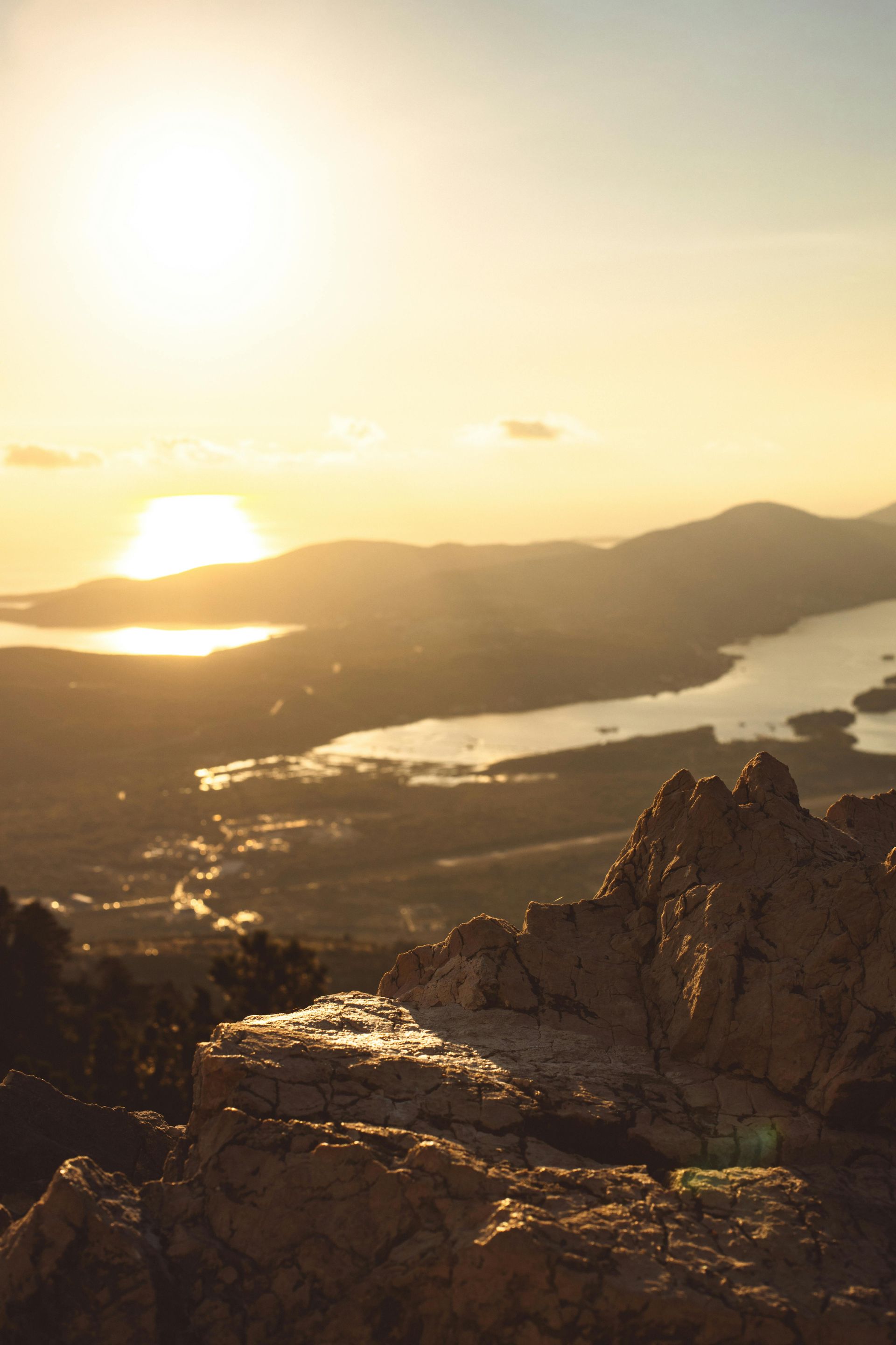 Sunset over mountains and a bay seen from a rocky mountaintop. Warm, golden light bathes the landscape.