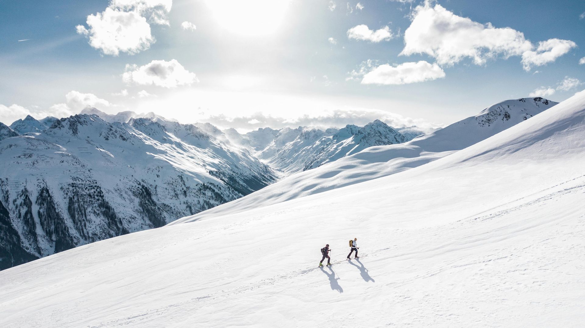 Two people ski touring uphill on a snowy mountain slope under a bright sun and blue sky with distant peaks.