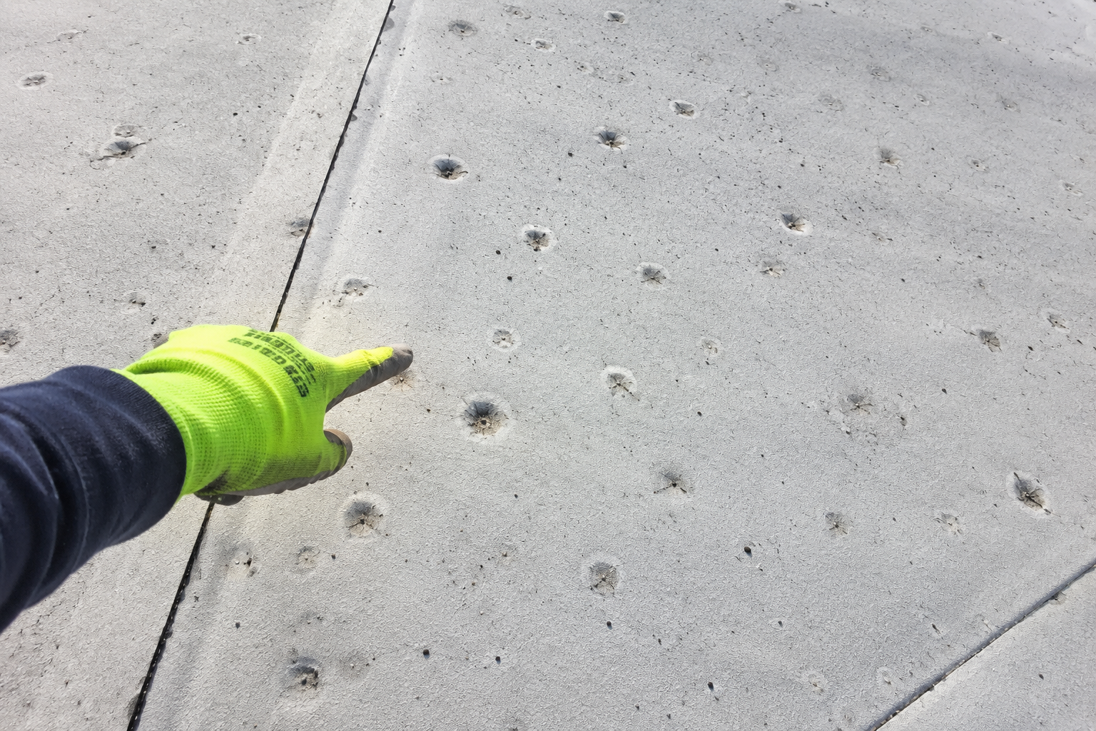 Gloved hand pointing at small holes in a concrete sidewalk near a curb
