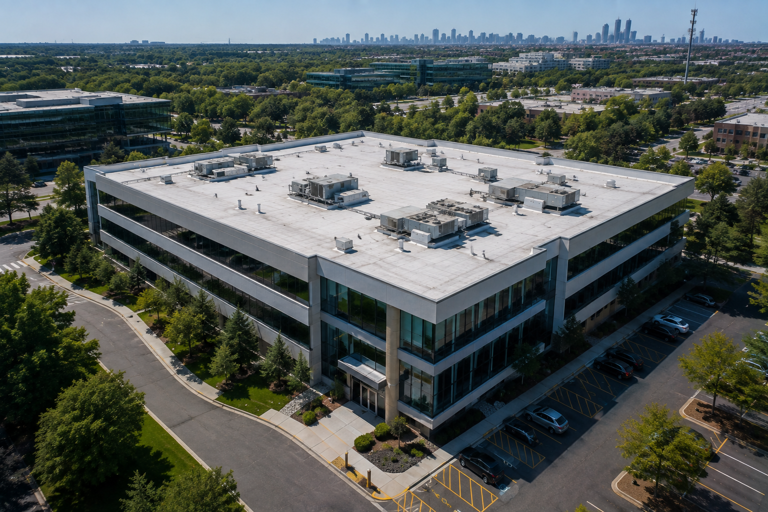 Aerial view of a modern office building with a flat white roof, surrounded by trees and roads, with a distant skyline.