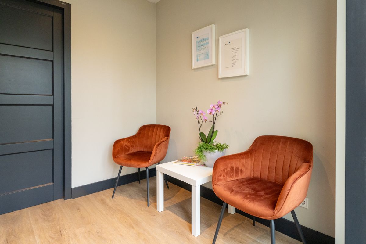 Twee oranje fluwelen stoelen staan ​​tegenover een kleine witte tafel met bloemen, onder een ingelijste kunstgalerij. 
