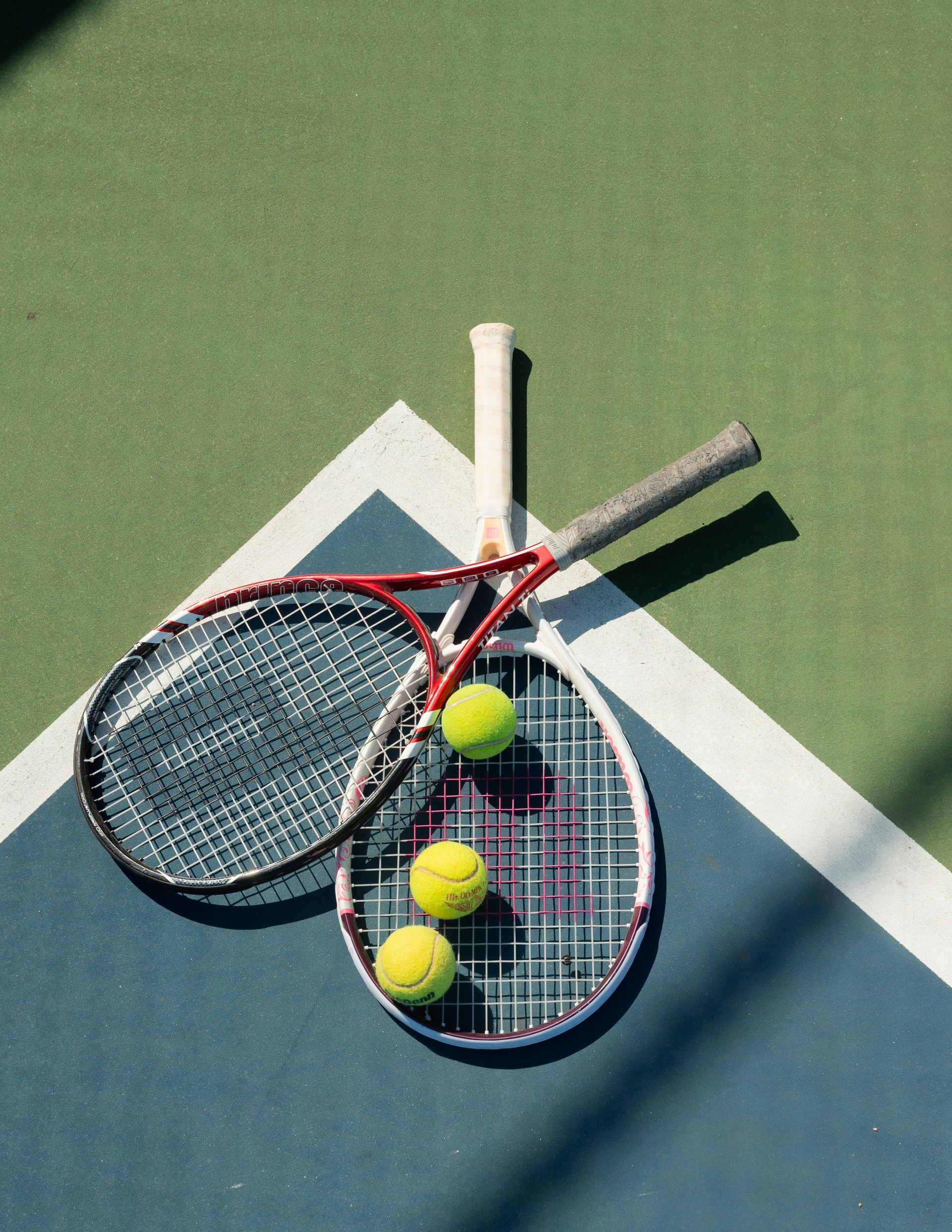 Tennisrackets en ballen op een tennisbaan. Twee rackets liggen gekruist, met daartussen drie gele ballen.