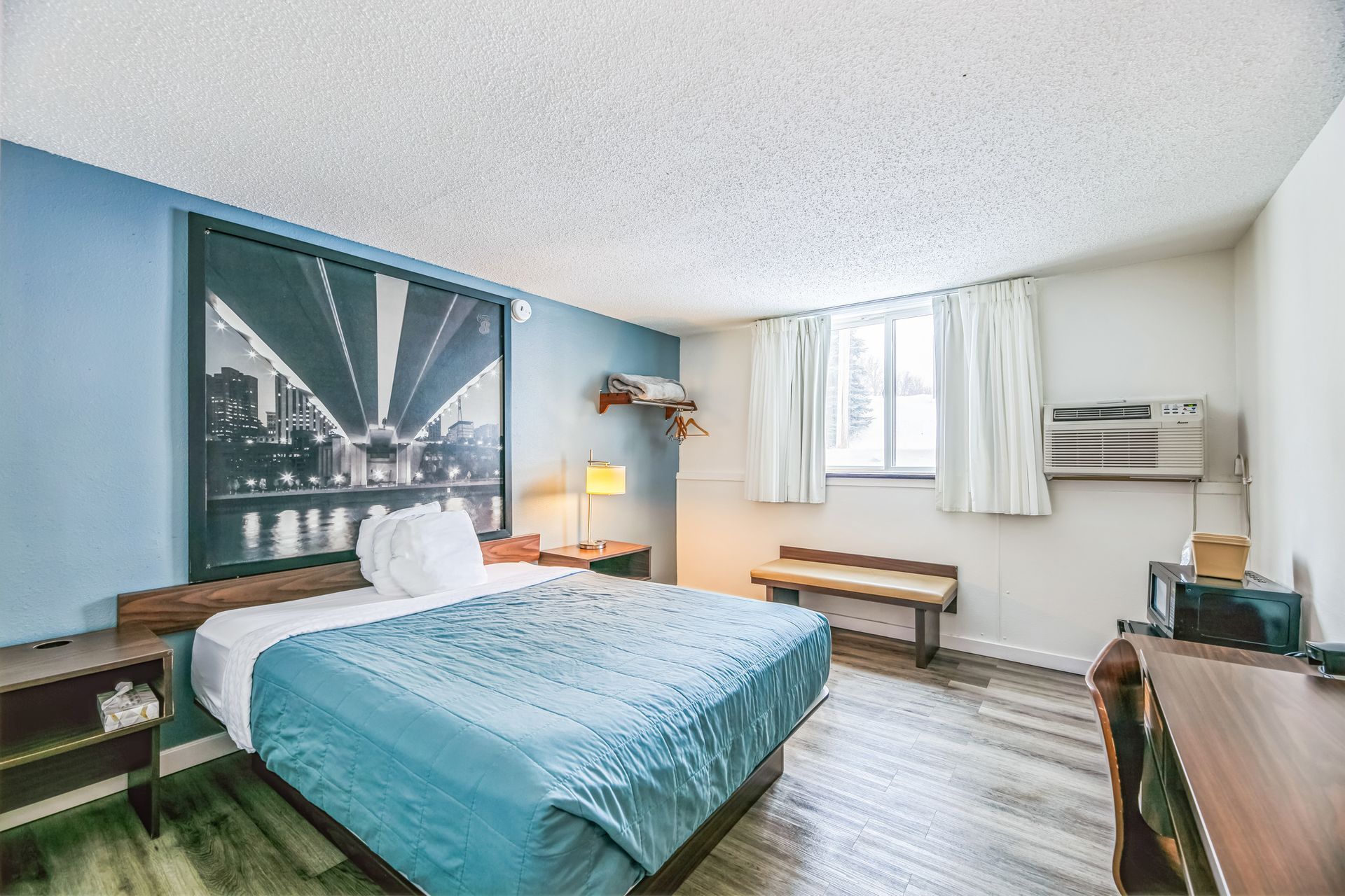 Motel room with bed, city art, and a fish decor on the wall. Window, bench, and microwave are visible.