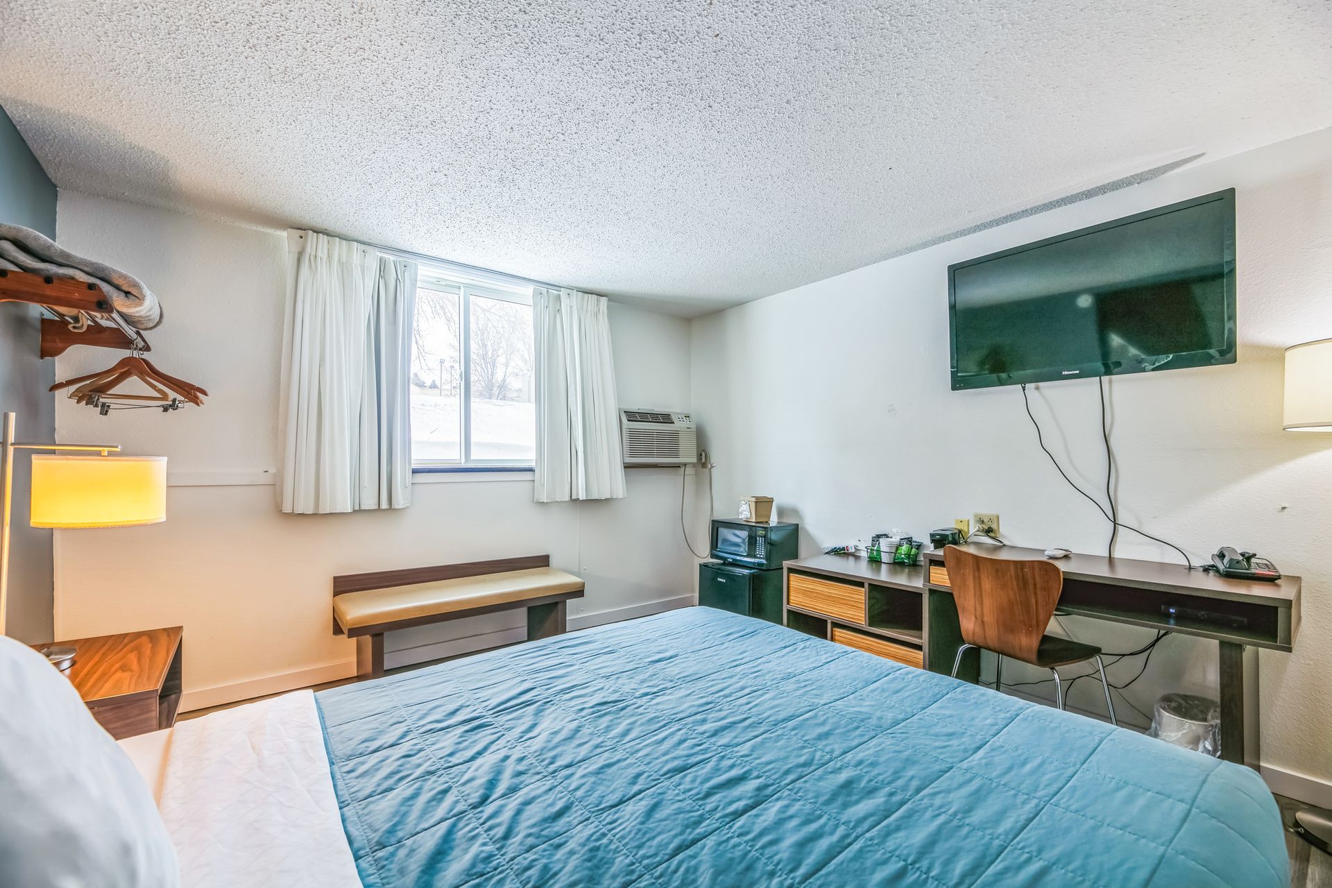 Hotel room with a bed, TV, desk, window, and small refrigerator. Walls are white and turquoise bedspread.