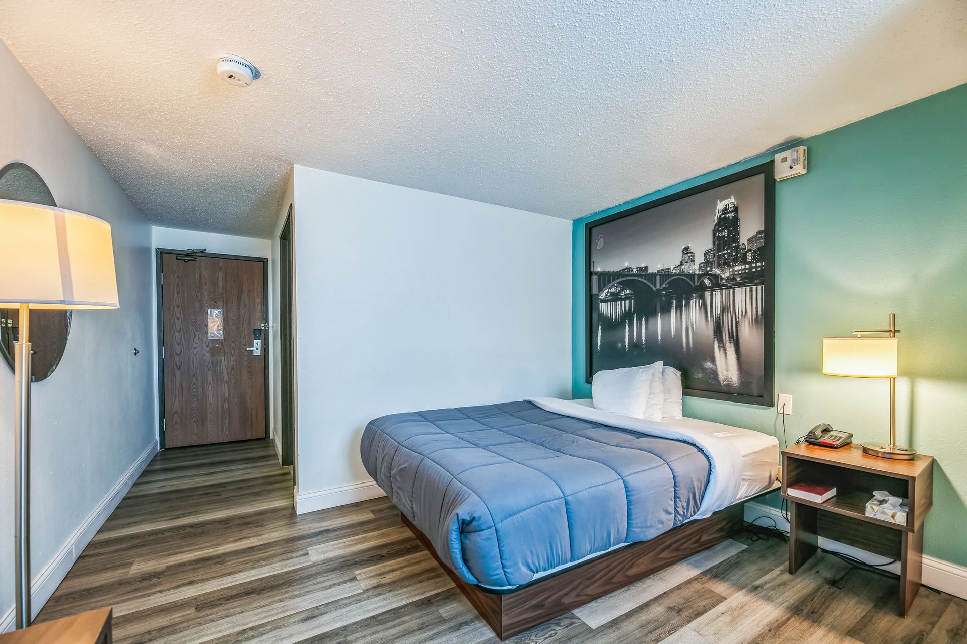 Hotel room with a bed, small nightstand, lamp, and cityscape photo on the wall.