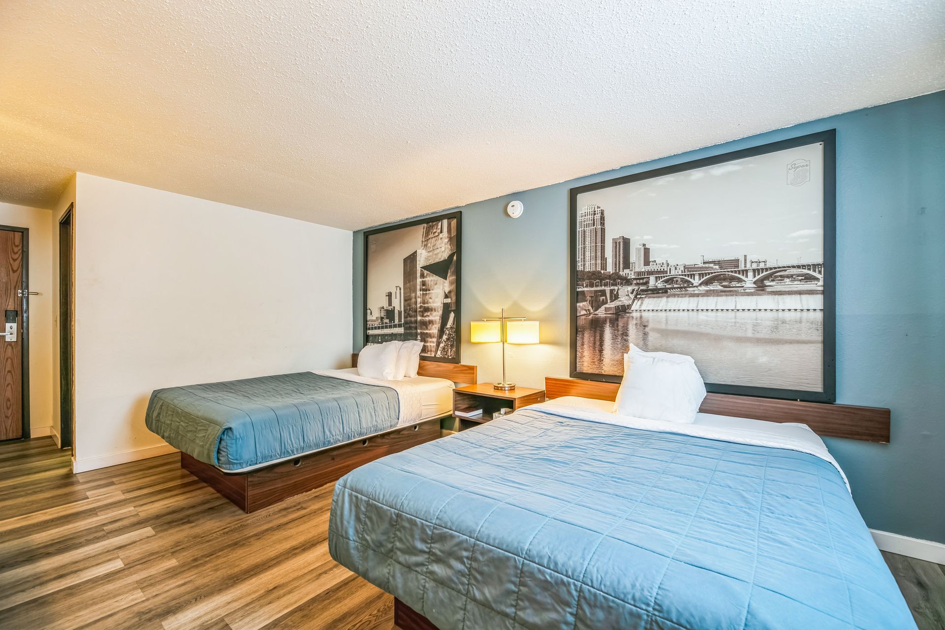 Two beds in a hotel room with light blue comforters, city skyline art, and wood floors.
