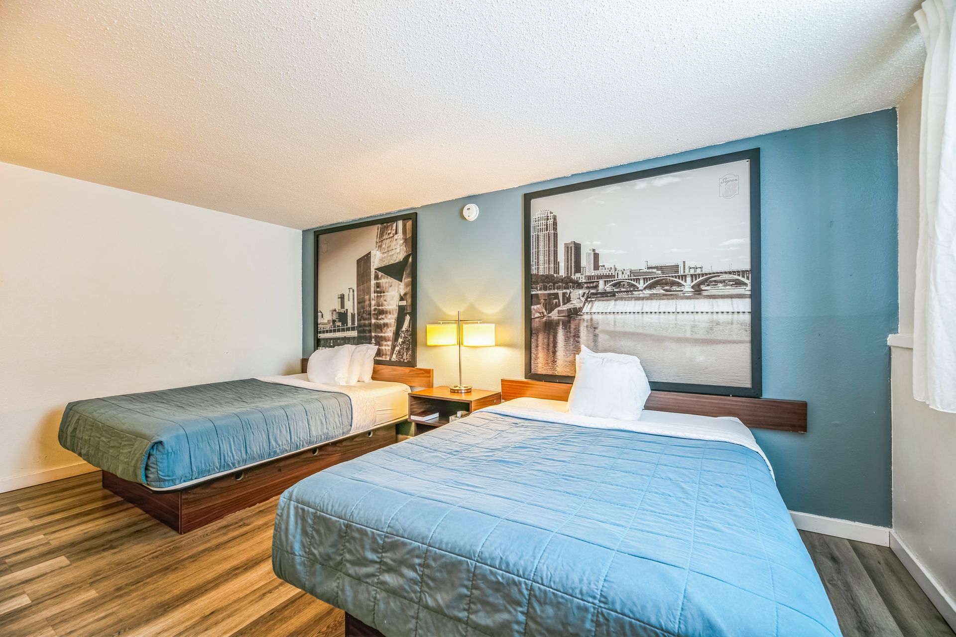 Two beds in a motel room with city skyline art above them. Blue bedspreads, wood-look floor.