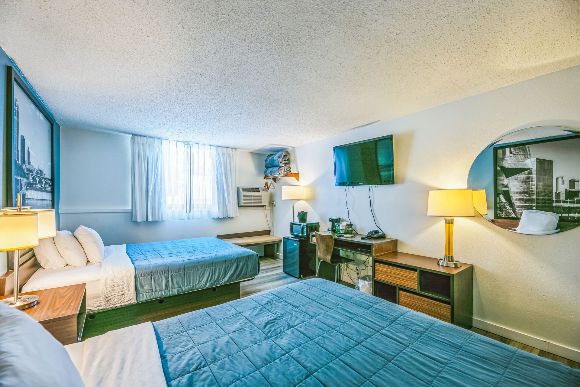 Hotel room with two beds, TV, desk, and round mirror. Blue and white color scheme.
