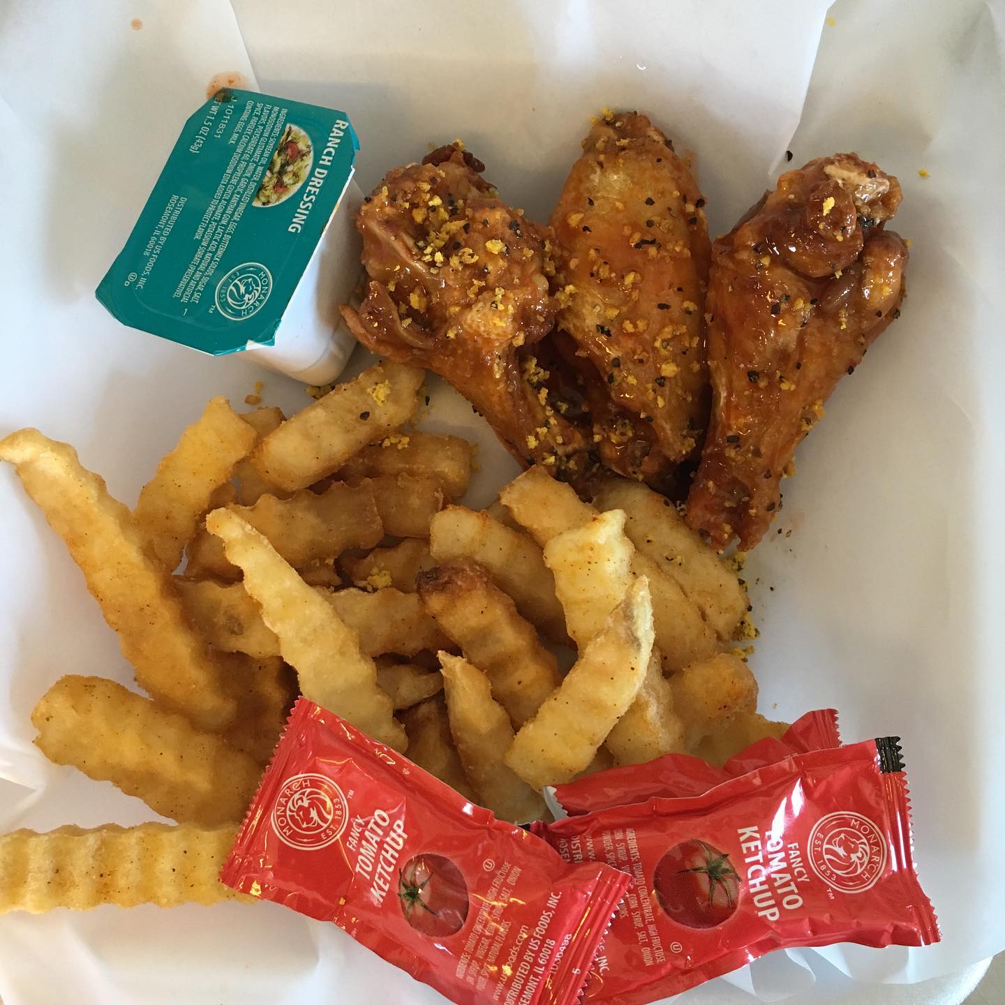 Chicken wings and fries in a white container, with ketchup packets and a sauce cup.