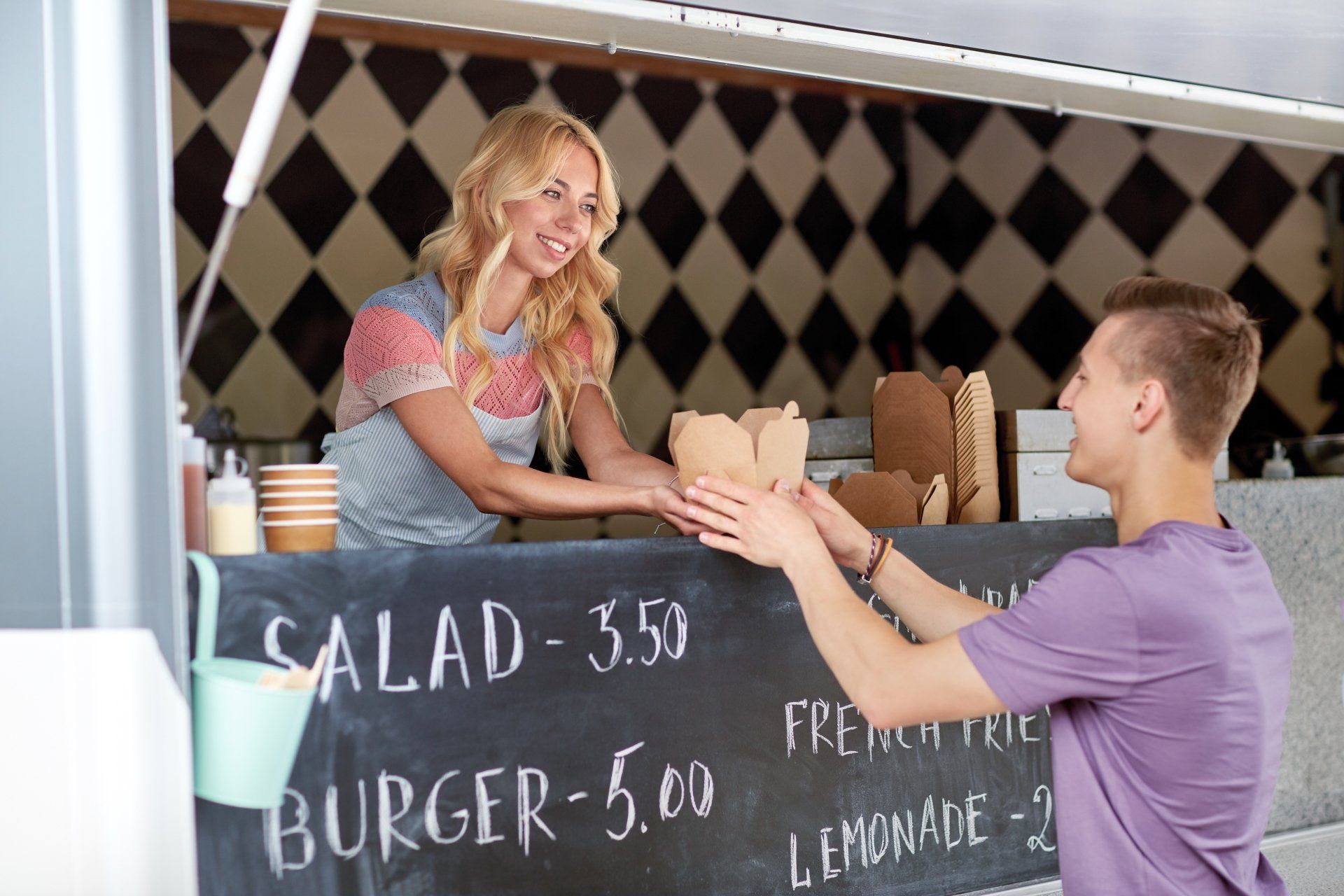 Woman in food truck hands order to man. Chalkboard menu visible.