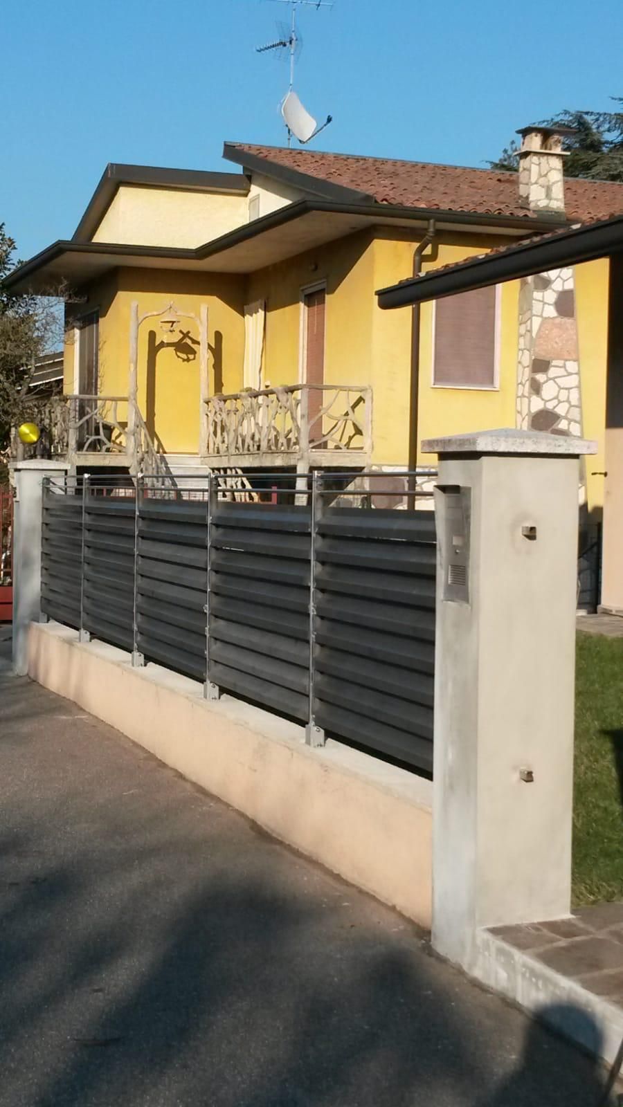 Una casa gialla dietro una moderna recinzione composta da listelli orizzontali grigi montati su una base di cemento e pilastri di pietra.