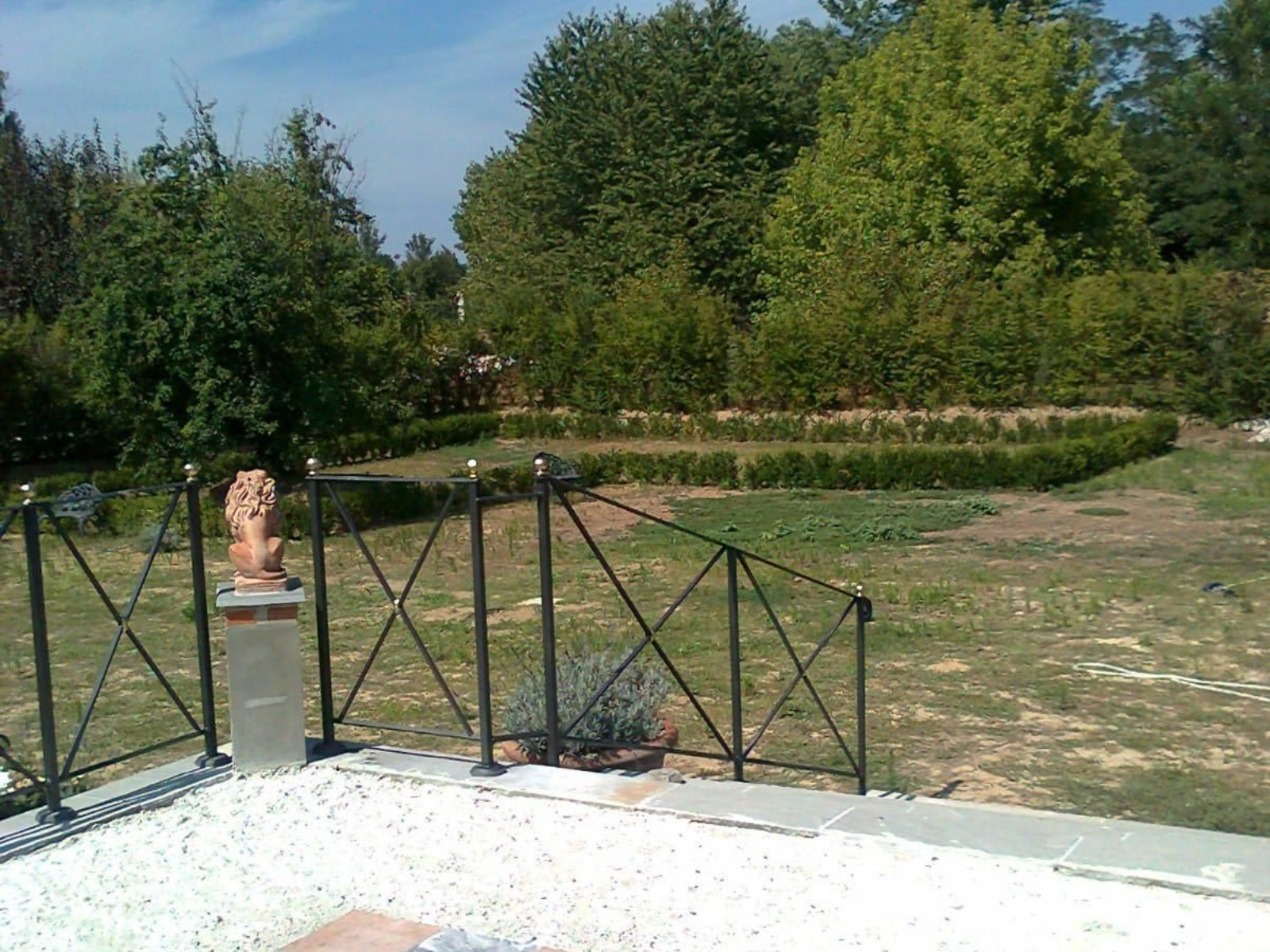 Un patio in pietra con ringhiera in metallo e una statua di leone che si affaccia su un cortile erboso con una bassa siepe.