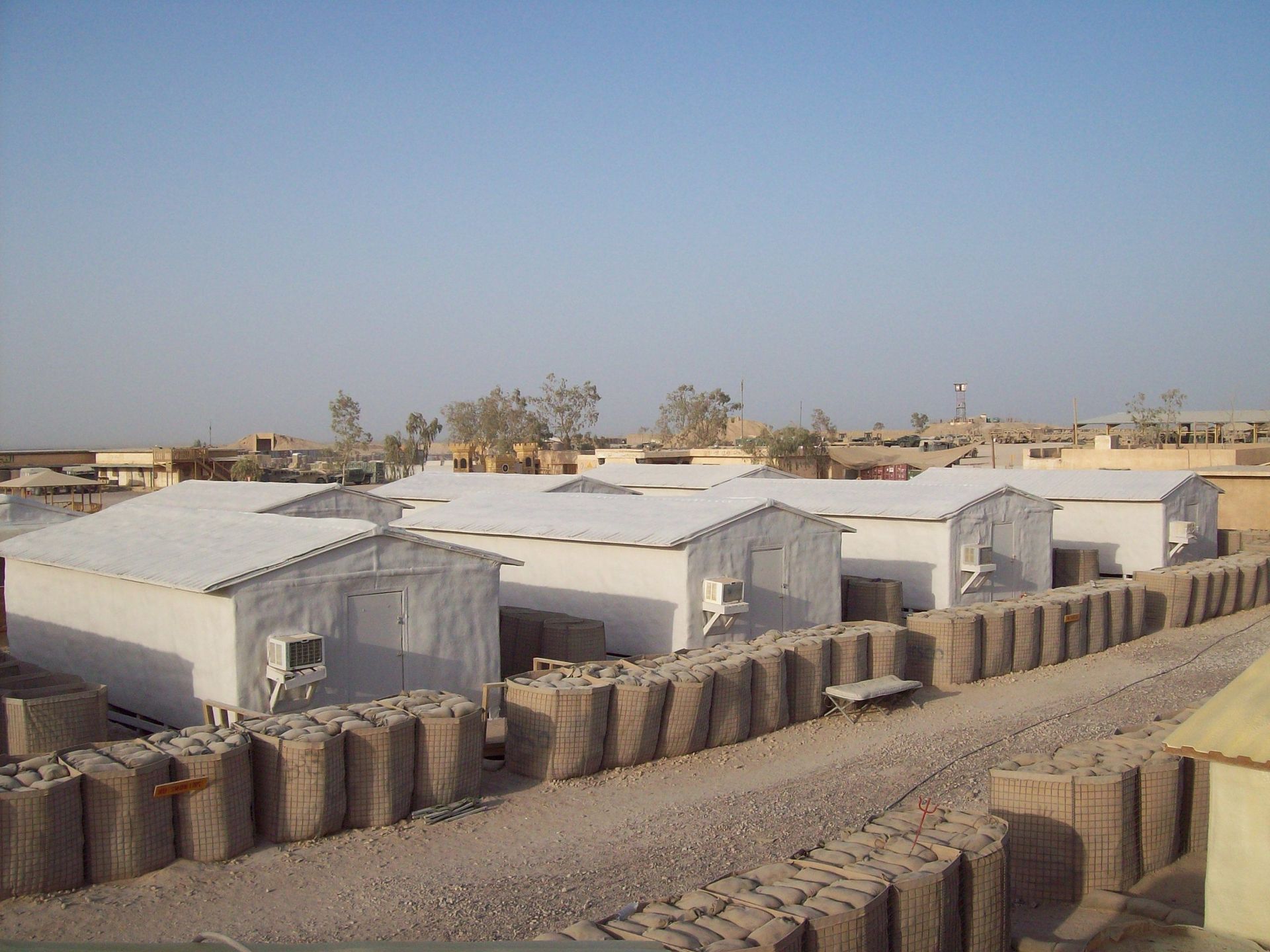 Rows of white tents in a dry camp, with sandbag barriers under a clear blue sky.
