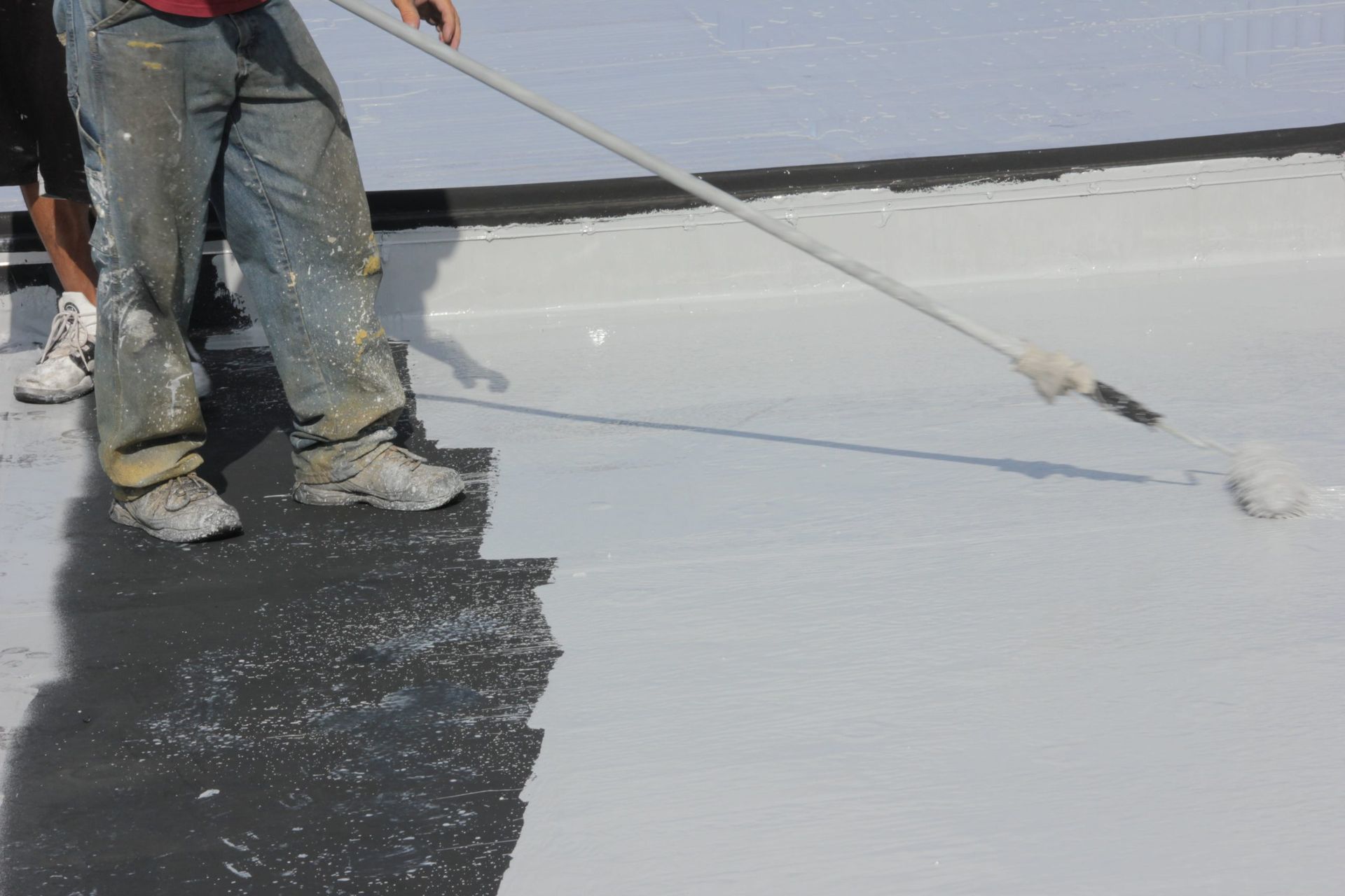 Worker spraying black coating on a flat roof beside a white surface