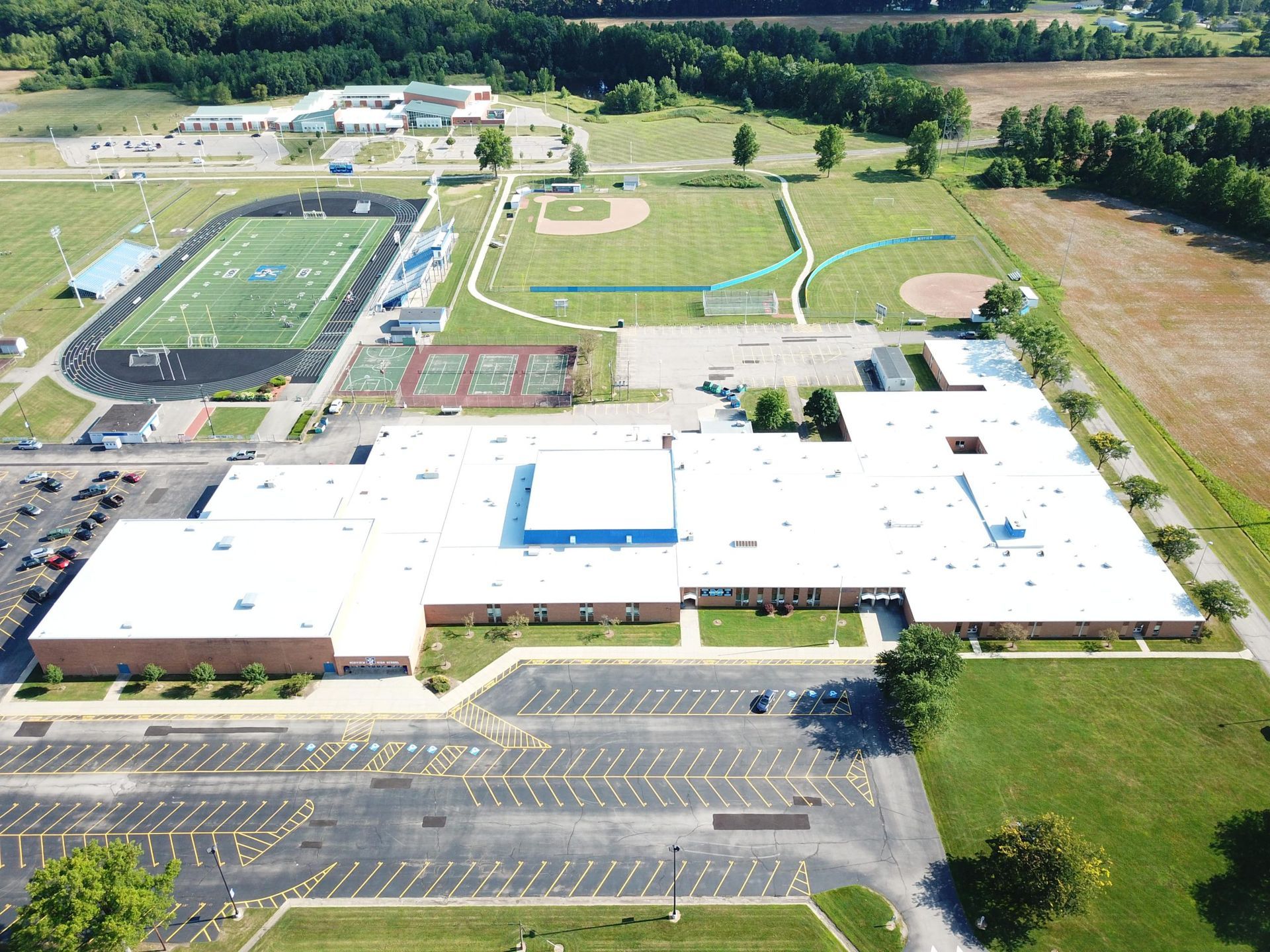 Aerial view of a school campus with sports fields, parking lot, and surrounding trees.